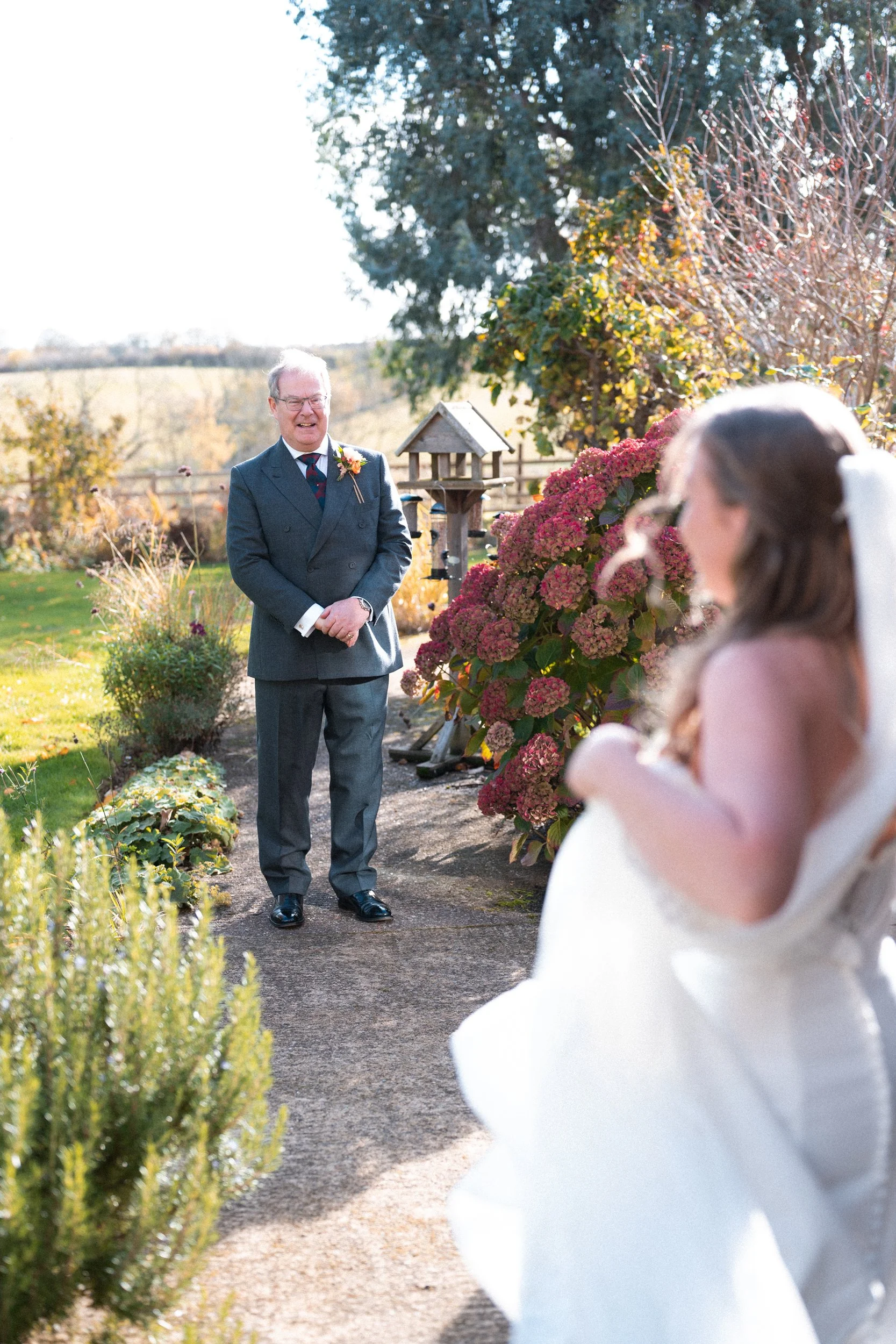 A man in a gray suit stands outdoors smiling, looking at a woman in a white wedding dress, with colorful autumn foliage and pink hydrangeas around them.
