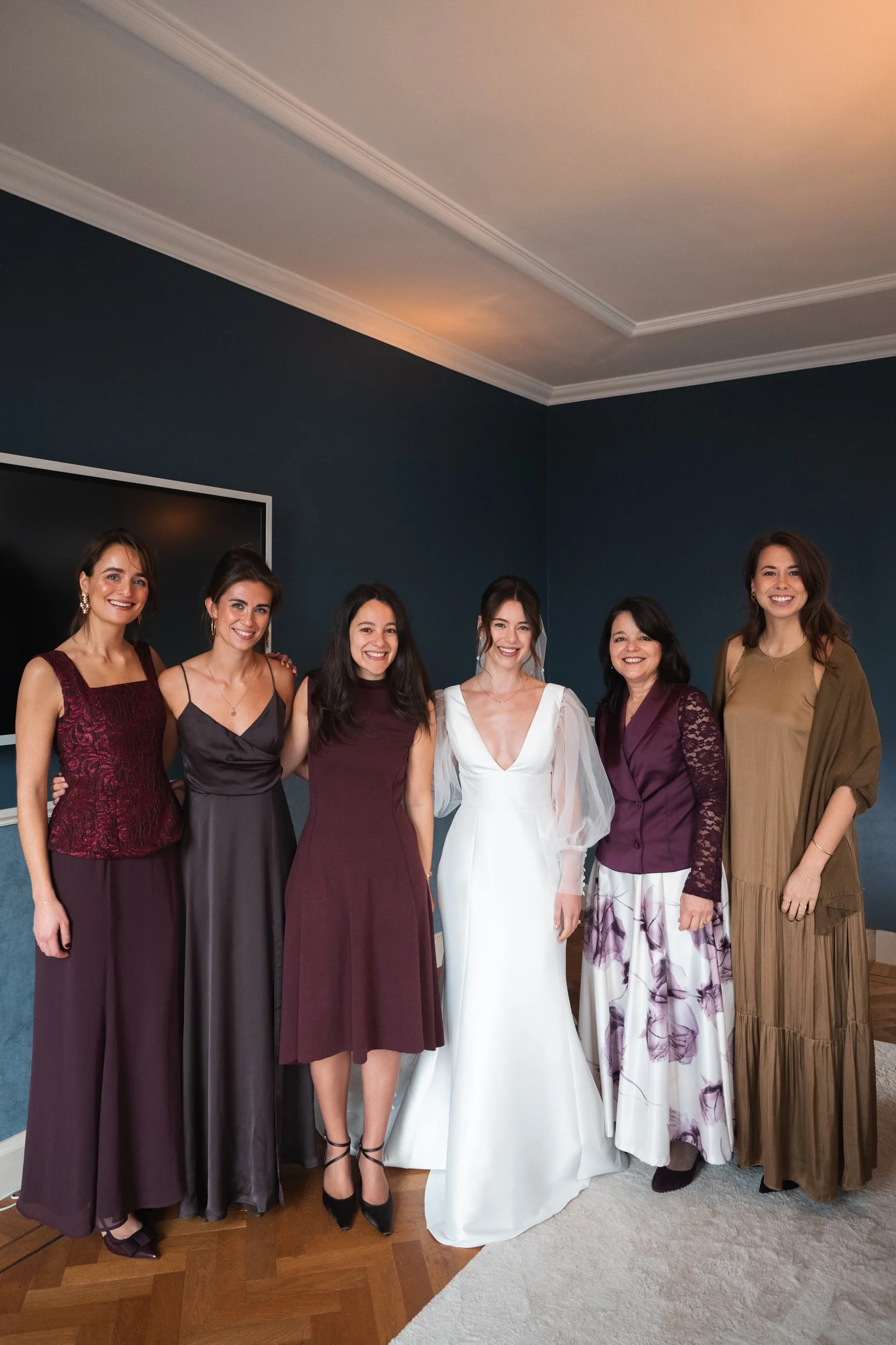 Group of seven women dressed in formal attire, standing together indoors against a dark blue wall, smiling.
