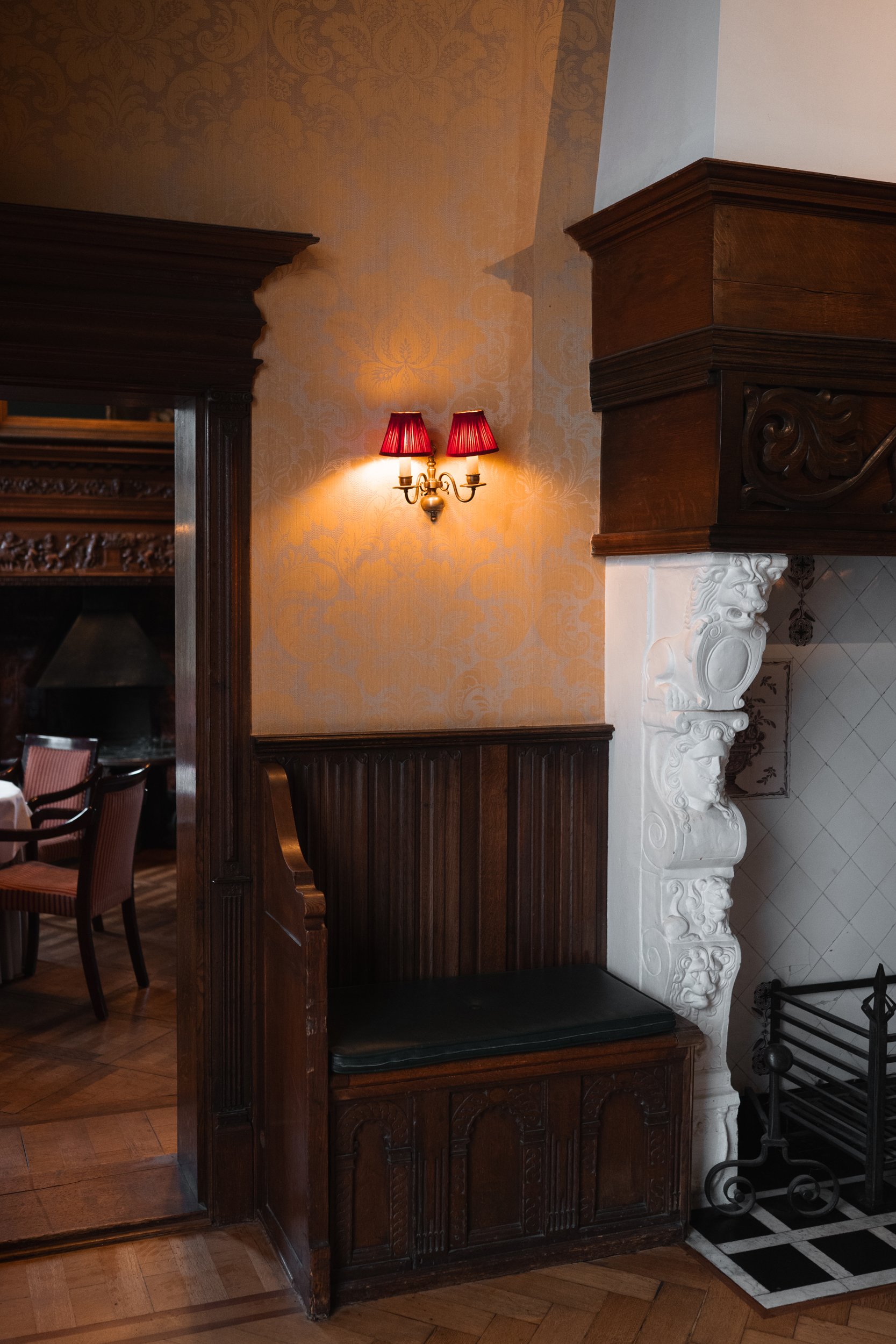 Elegant interior with a floral-patterned wallpaper, a wall-mounted red lampshade sconce, dark wood wainscoting, a wooden bench with a black cushioned seat, and a decorative white carved fireplace wall with a metal firewood holder.