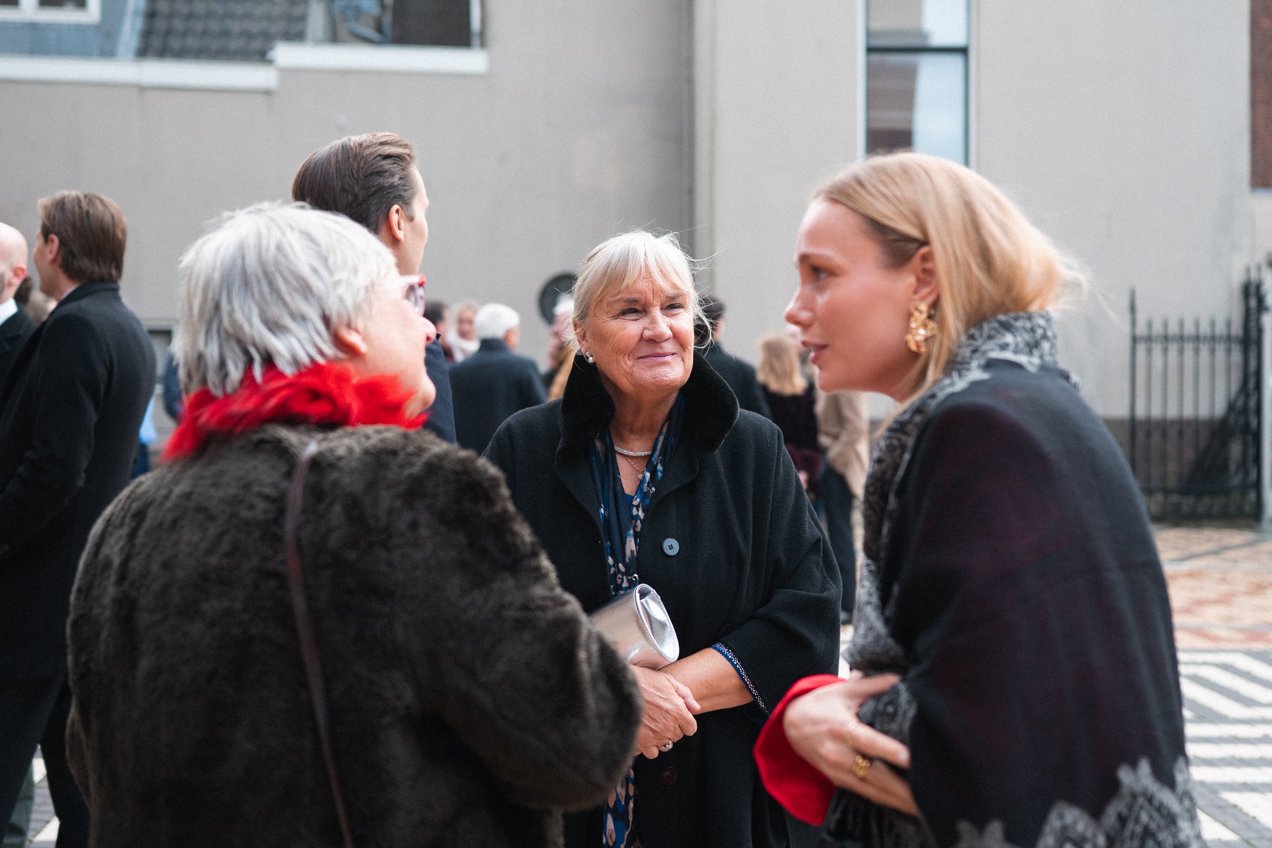 Three women in conversation at an outdoor event, with other people in the background.