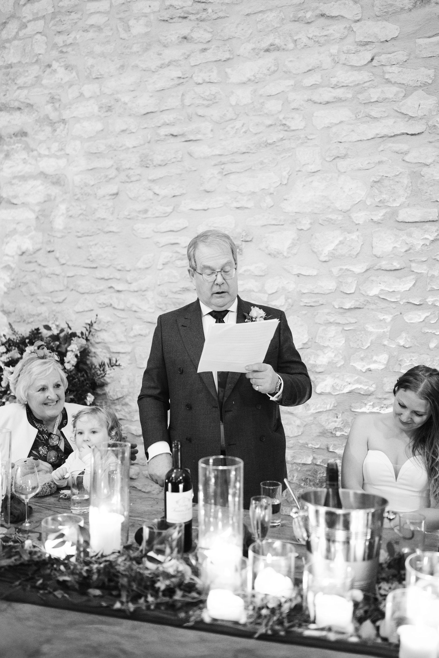 A black and white photo of a man giving a speech at a wedding reception, surrounded by women and children seated at a table with candles, wine, and floral decorations, against a textured stone wall background.