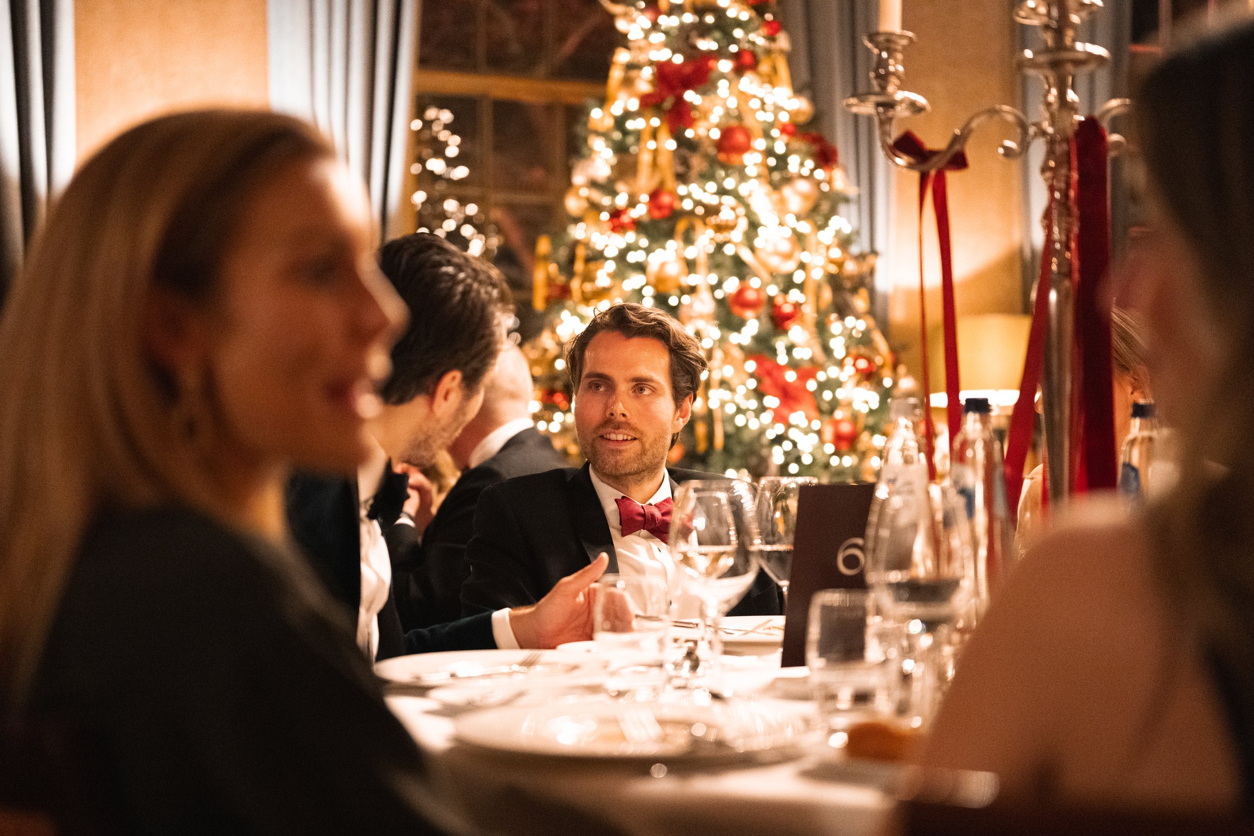 People sitting at a dinner table during a Christmas celebration, with a decorated Christmas tree in the background.