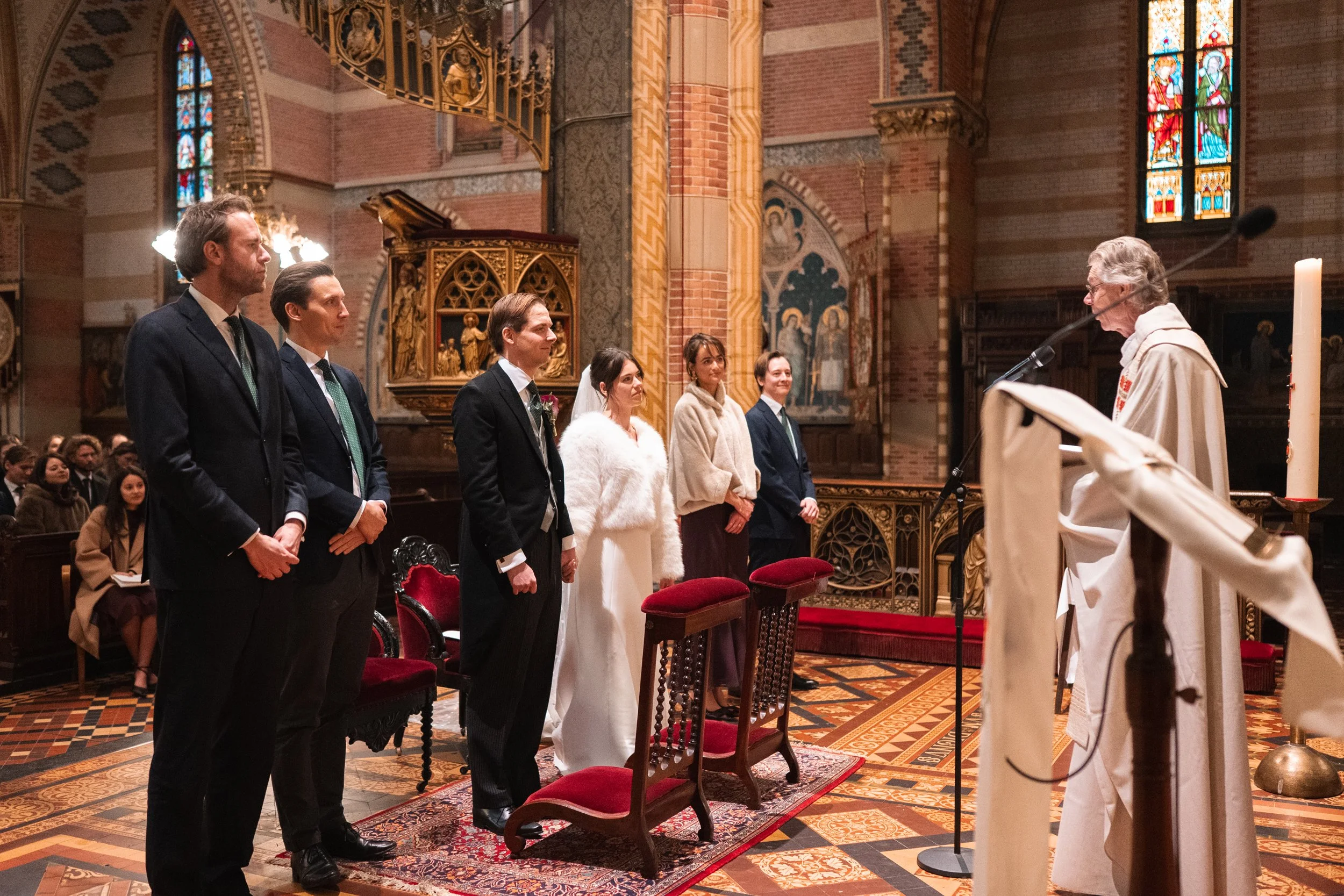 A wedding ceremony inside a church with six people standing in front of a priest. The bride is dressed in white, with a veil and fur coat, and the groom and five others are in formal suits. The church features stained glass windows, ornate woodwork, 