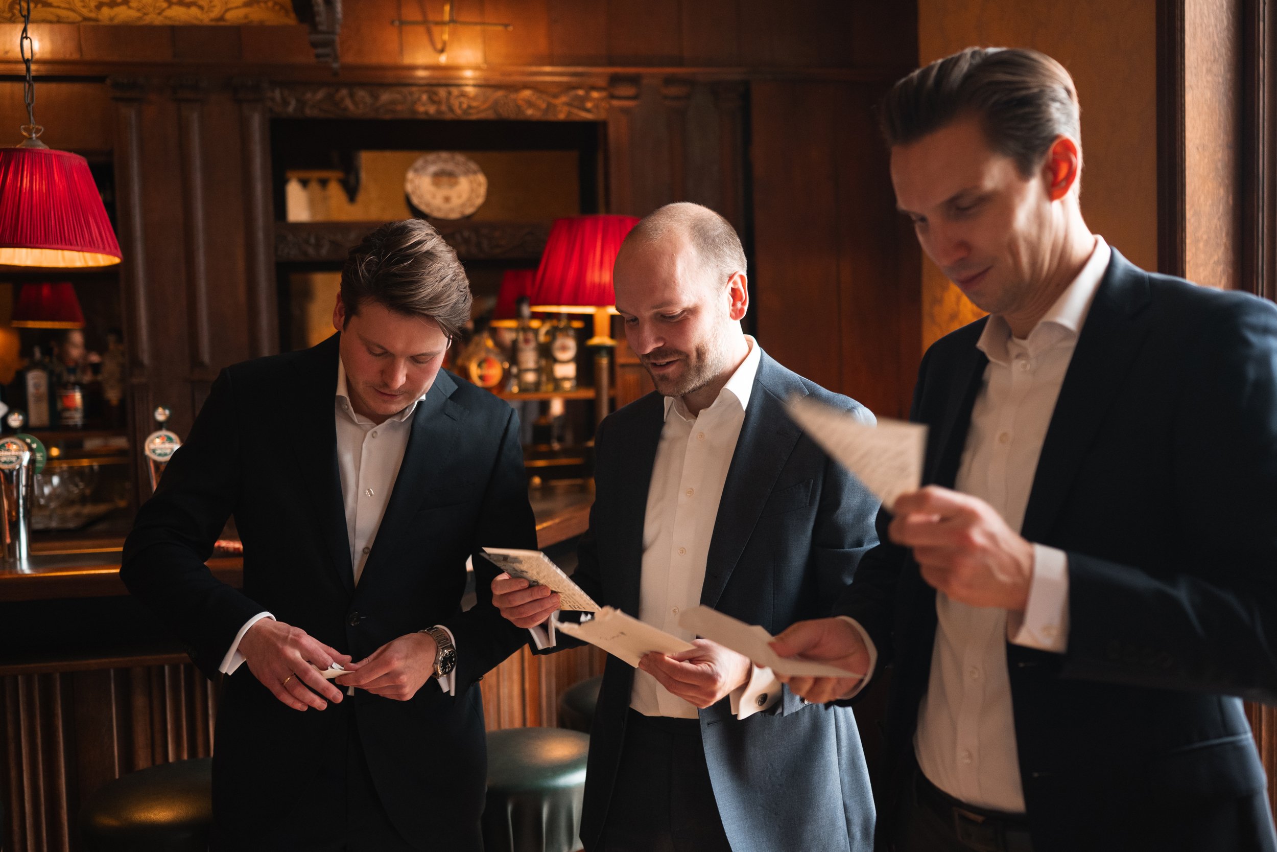 Three men in suits are standing at a bar in a wood-paneled room, looking at menus or notes.