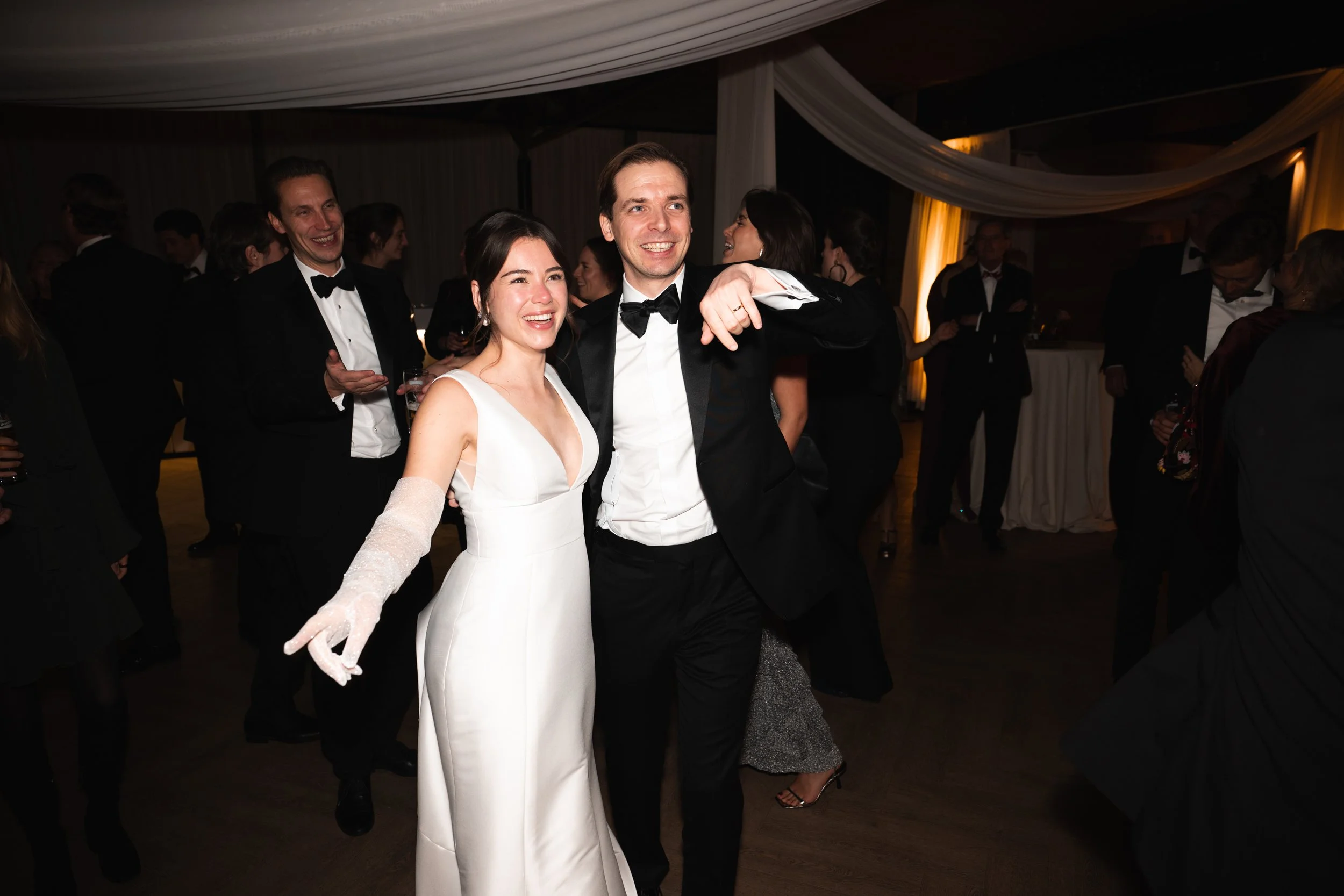 Couple dancing at a formal event, woman in white gown with gloves, man in black tuxedo, smiling and enjoying the music, surrounded by other people in tuxedos and evening dresses.