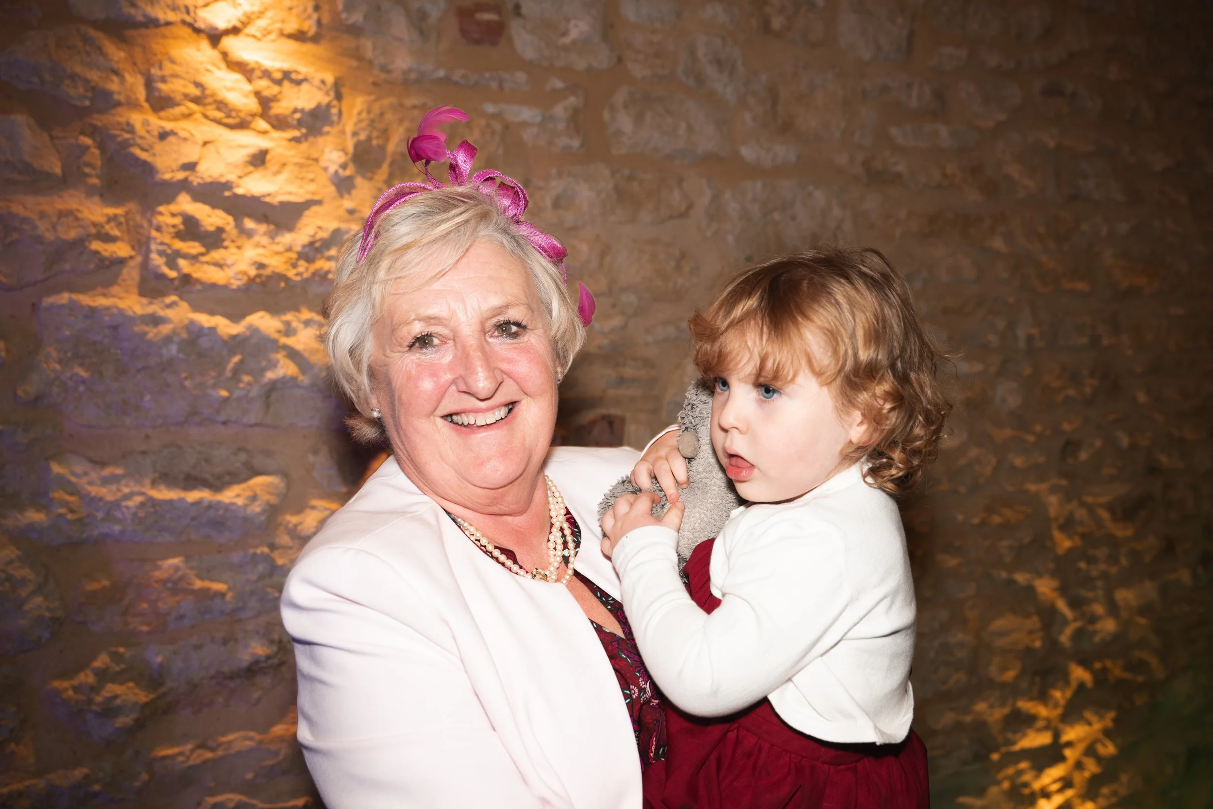 An elderly woman with short gray hair, wearing a pink fascinator hat and a white blazer, holding a young girl with curly red hair in a white top and red skirt against a brick wall background.