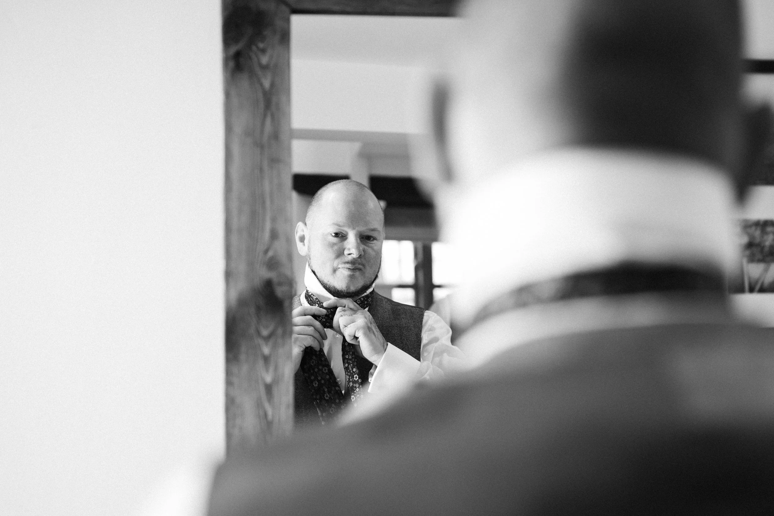 A man in a vest and shirt adjusts his tie while looking at himself in a mirror, with another person's blurred shoulder in the foreground.