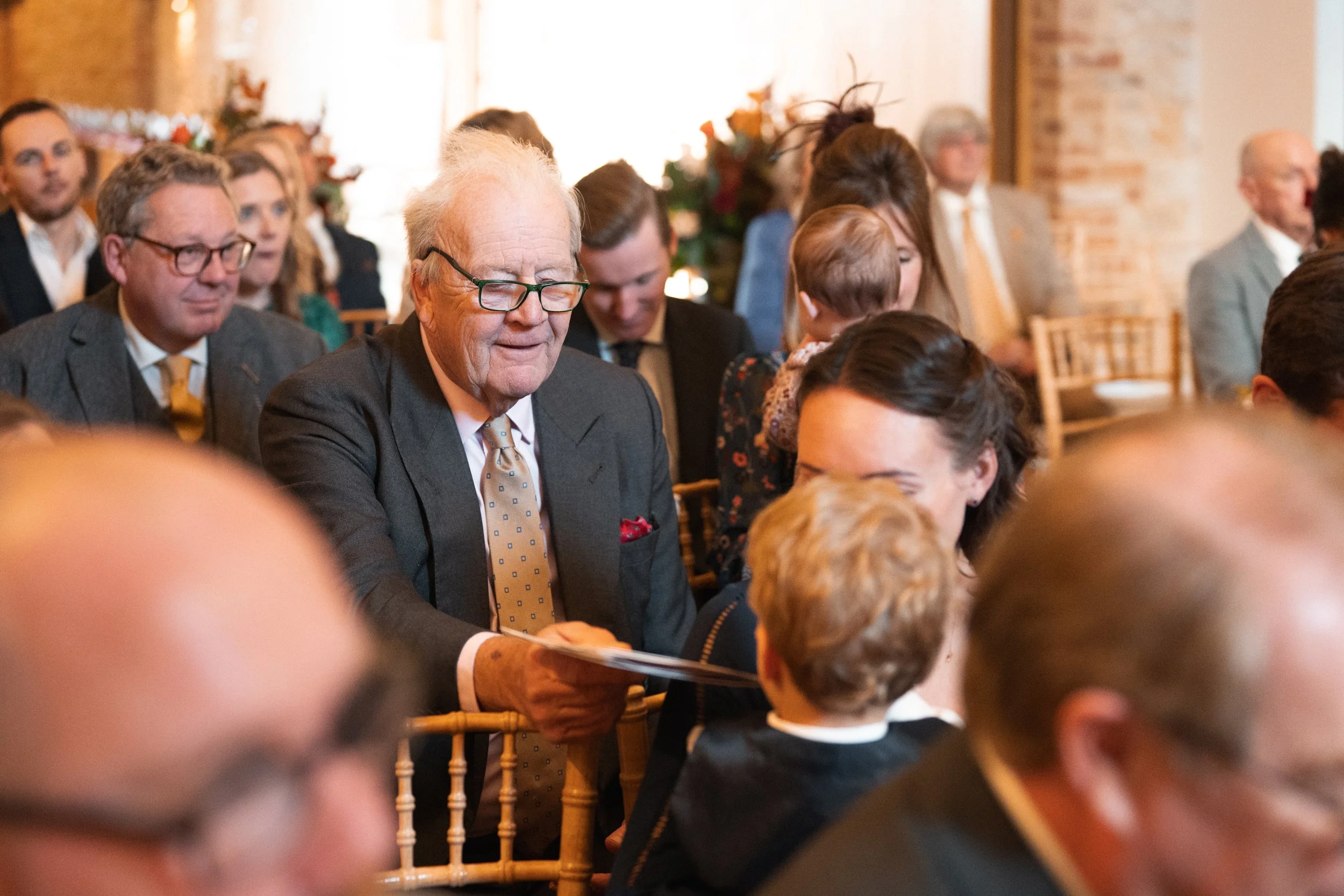 A group of people dressed in formal attire attending a gathering or ceremony in a warmly lit room with brick walls and floral decorations.