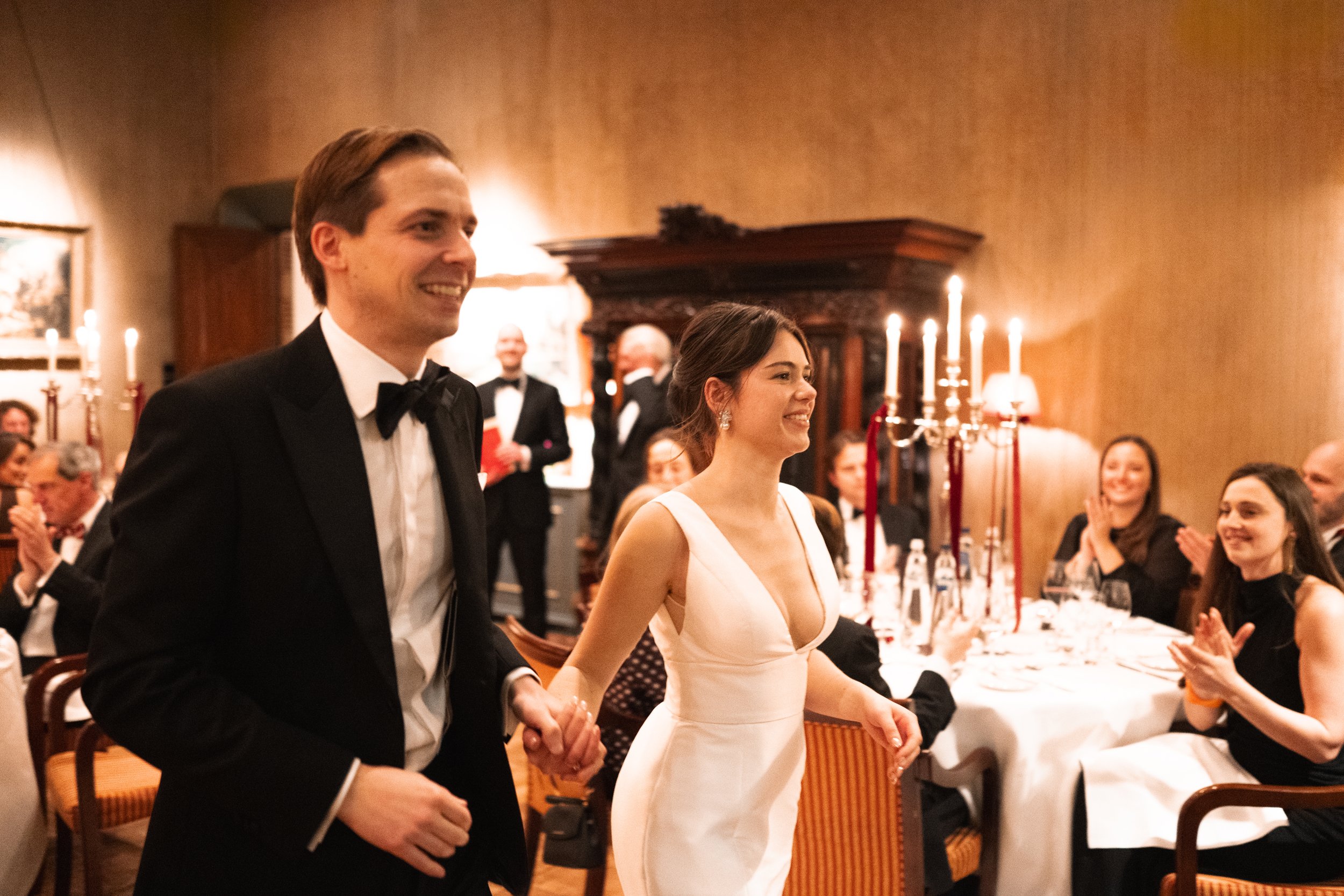 A couple, a man in a tuxedo and a woman in a white dress, holding hands and smiling at a formal dinner event with guests clapping, in an elegant room with candelabras and warm lighting.