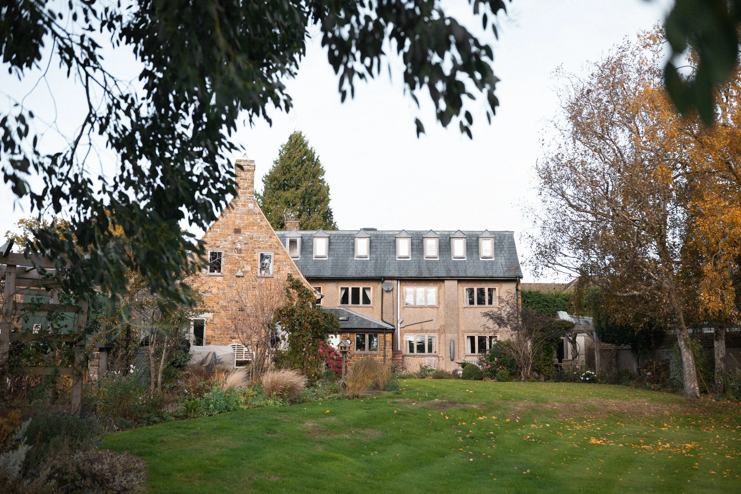 A backyard view of a large house with multiple windows, surrounded by trees and a well-maintained lawn with fallen leaves, taken during autumn.