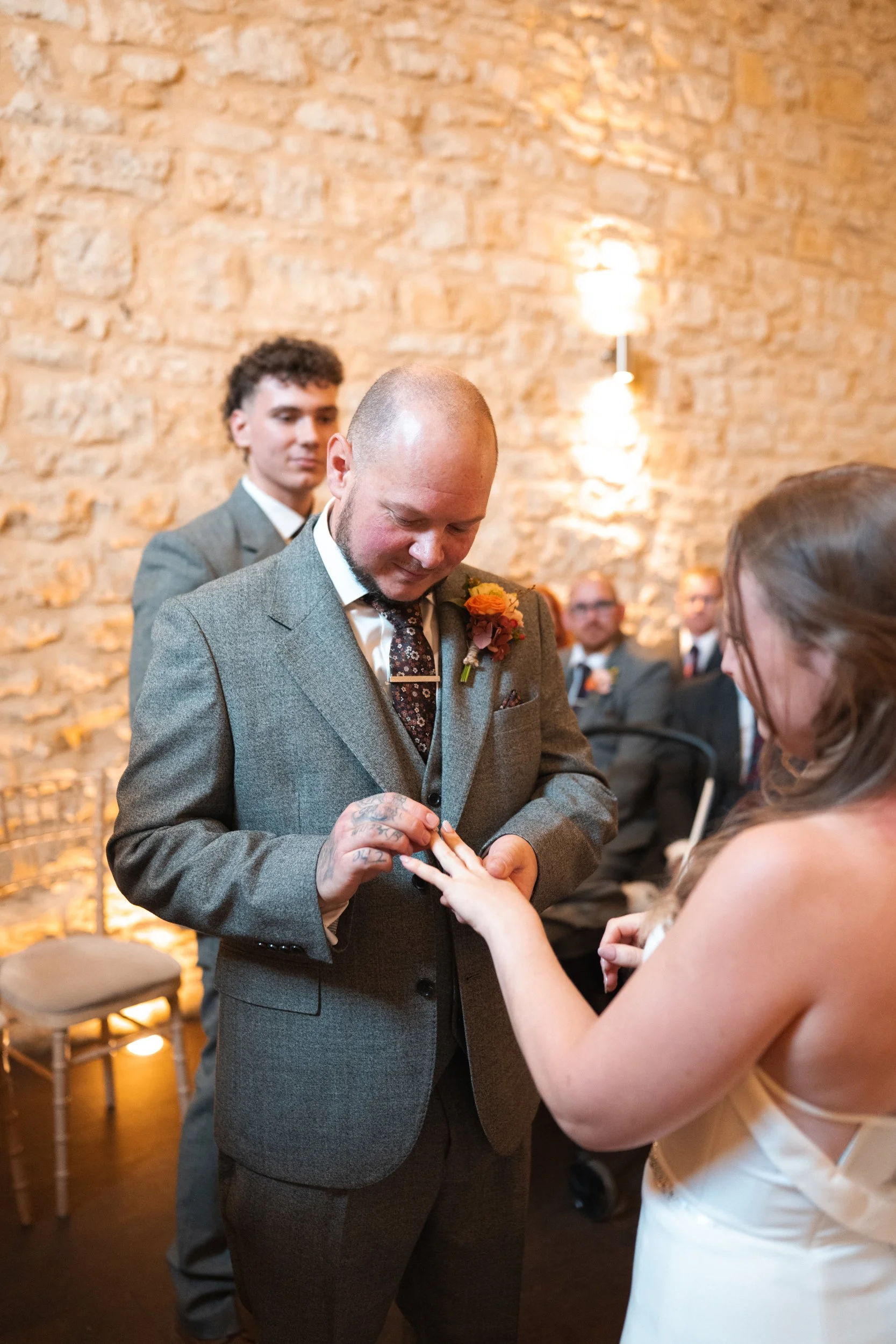 Groom placing ring on bride's finger during wedding ceremony in a rustic venue with exposed brick wall, attended by guests in formal attire.