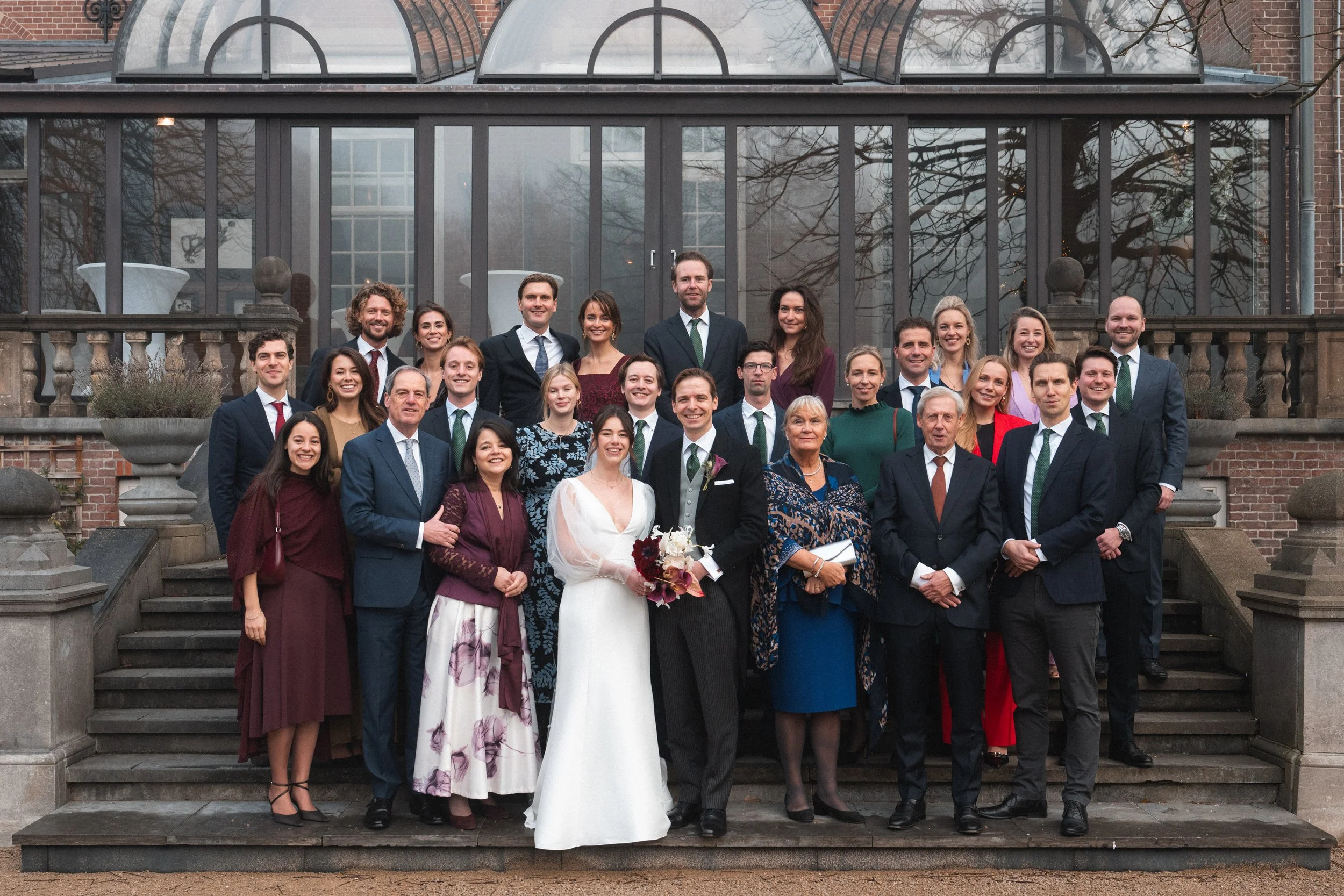 A group of people in formal attire posing on outdoor stairs for a wedding photo, with a glass building in the background.