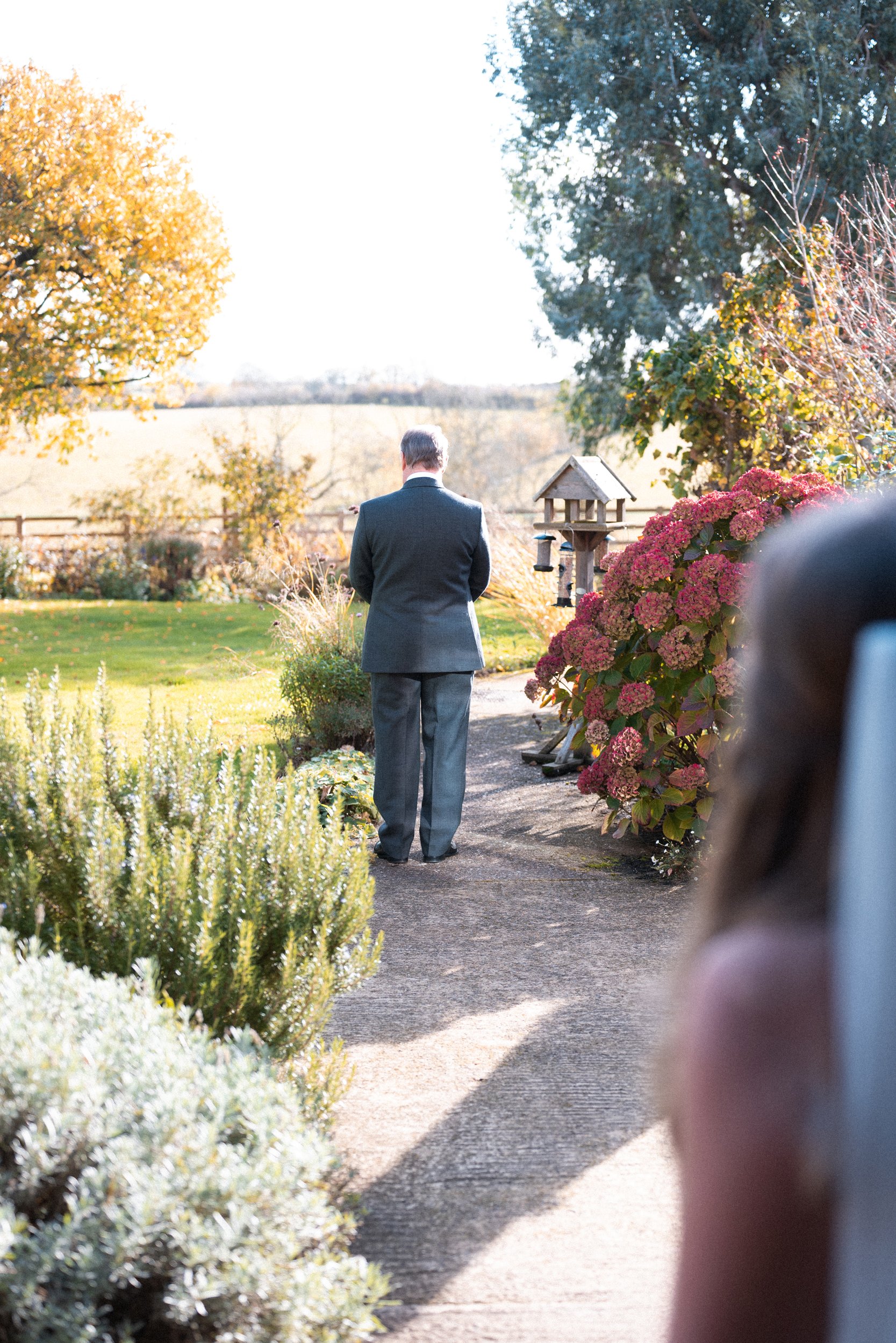 Man in a suit walking along a garden path with autumn trees and flowers, with a person and a house in the background.