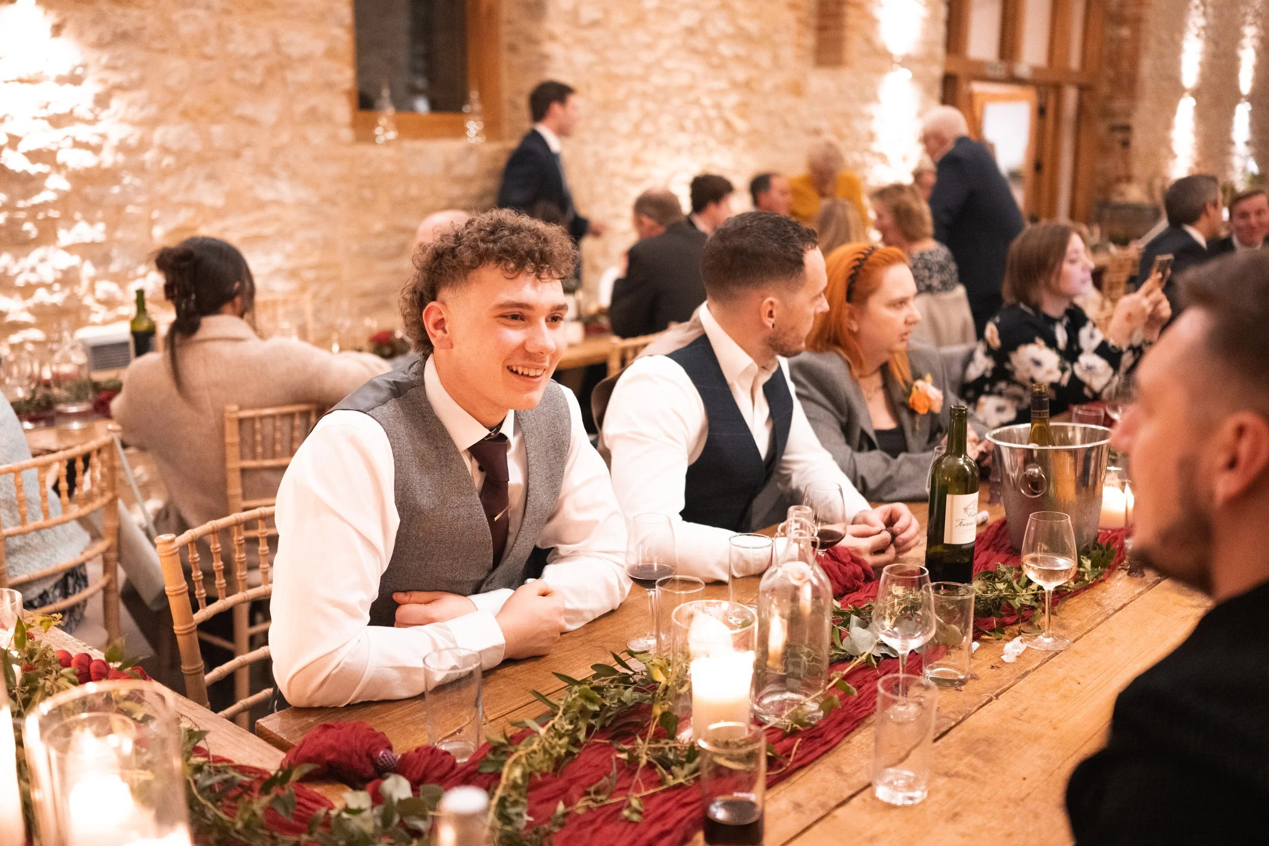 People sitting at a decorated wooden table during a celebration or dinner, with candles and wine bottles inside a rustic venue with brick walls.