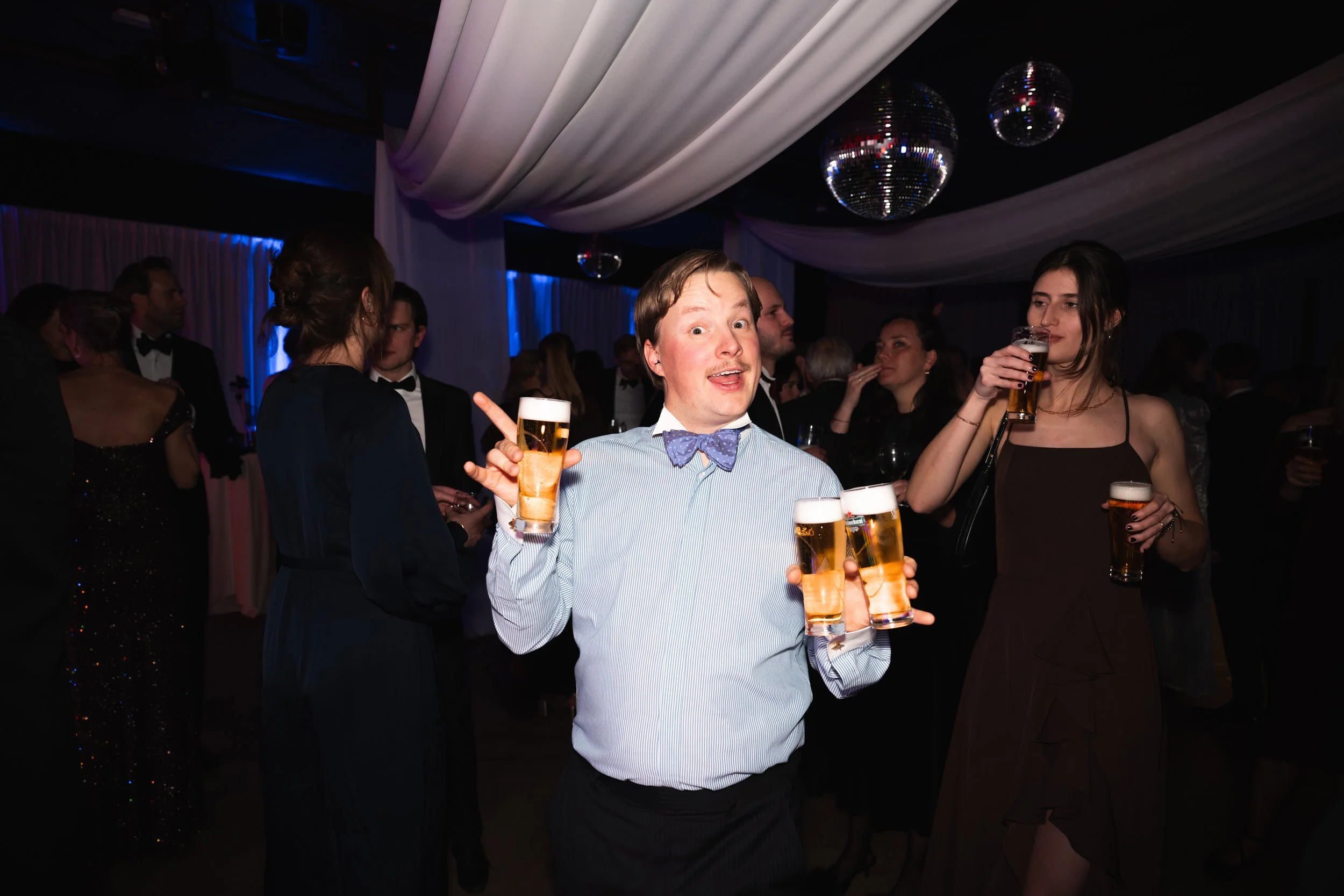 A man in a light blue shirt with a bow tie smiling and holding four glasses of beer at a party. Other guests are in the background, some holding drinks, in a room with draped ceiling and disco balls.