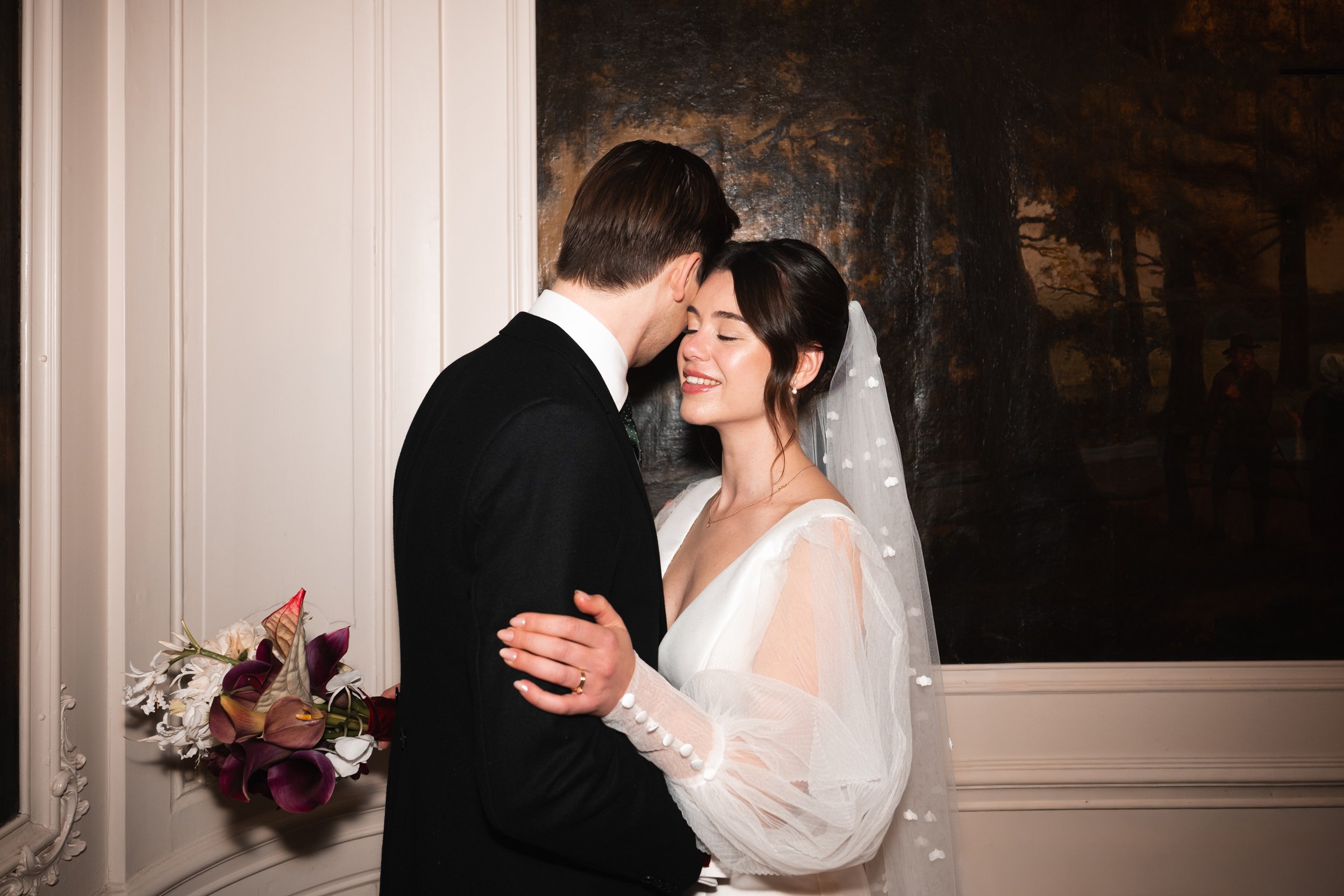 A bride and groom sharing a romantic moment indoors, standing close with their foreheads touching and eyes closed, with floral arrangements nearby.