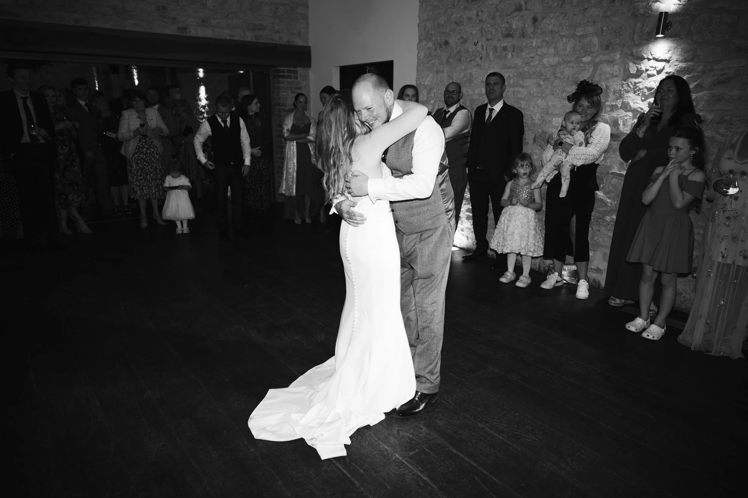 A bride and groom are sharing their first dance at a wedding reception, surrounded by wedding guests watching.