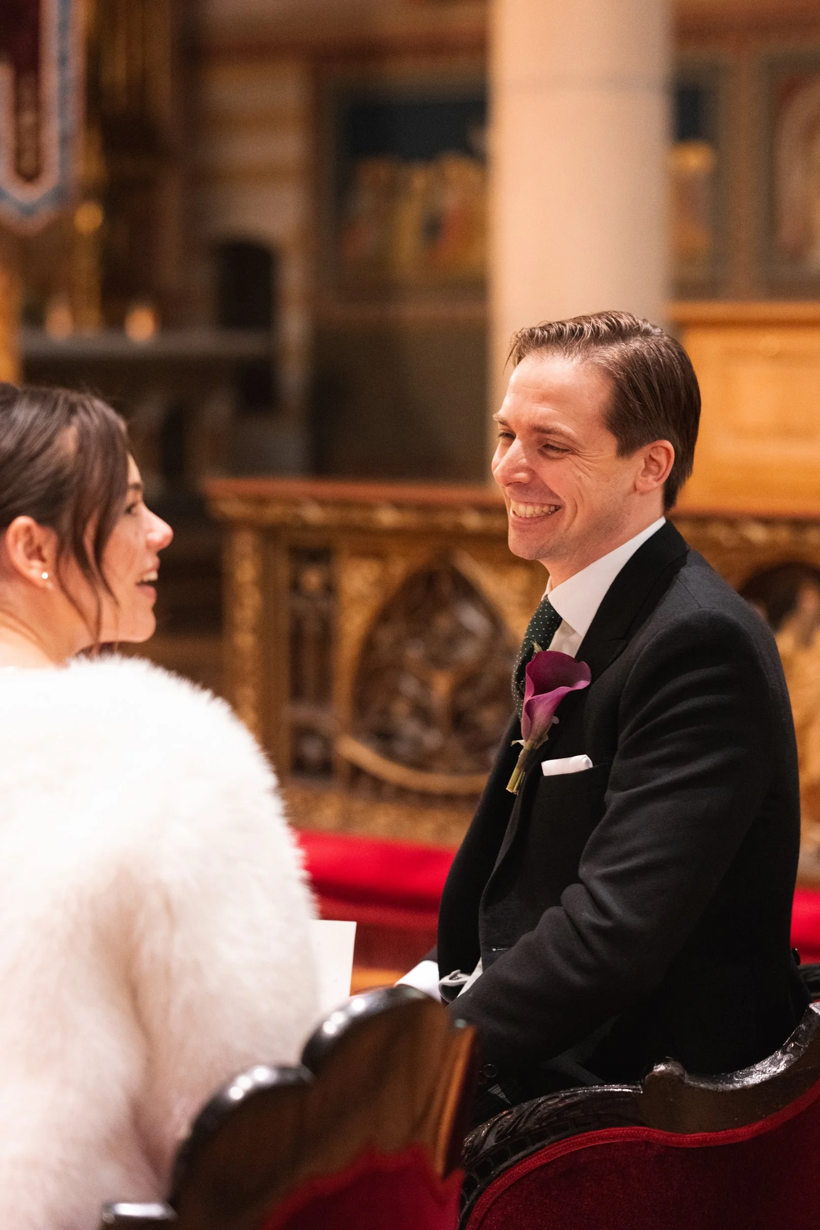 A man in a tuxedo with a boutonnière smiling at a woman in a white fluffy wrap inside a decorated church.