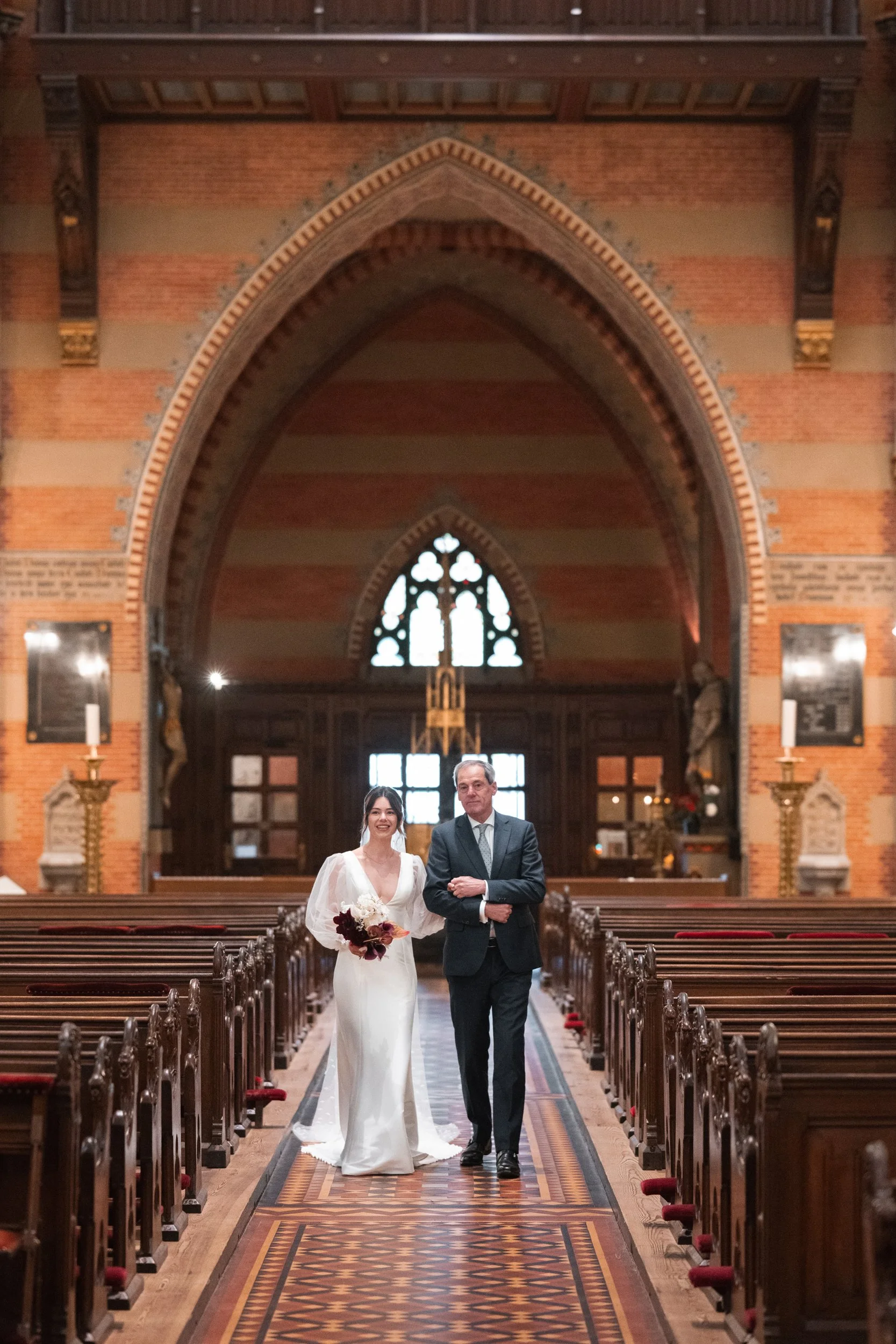 A bride in a white wedding gown holding a bouquet and a man in a dark suit walking down the aisle in a church with brick walls and stained glass windows.