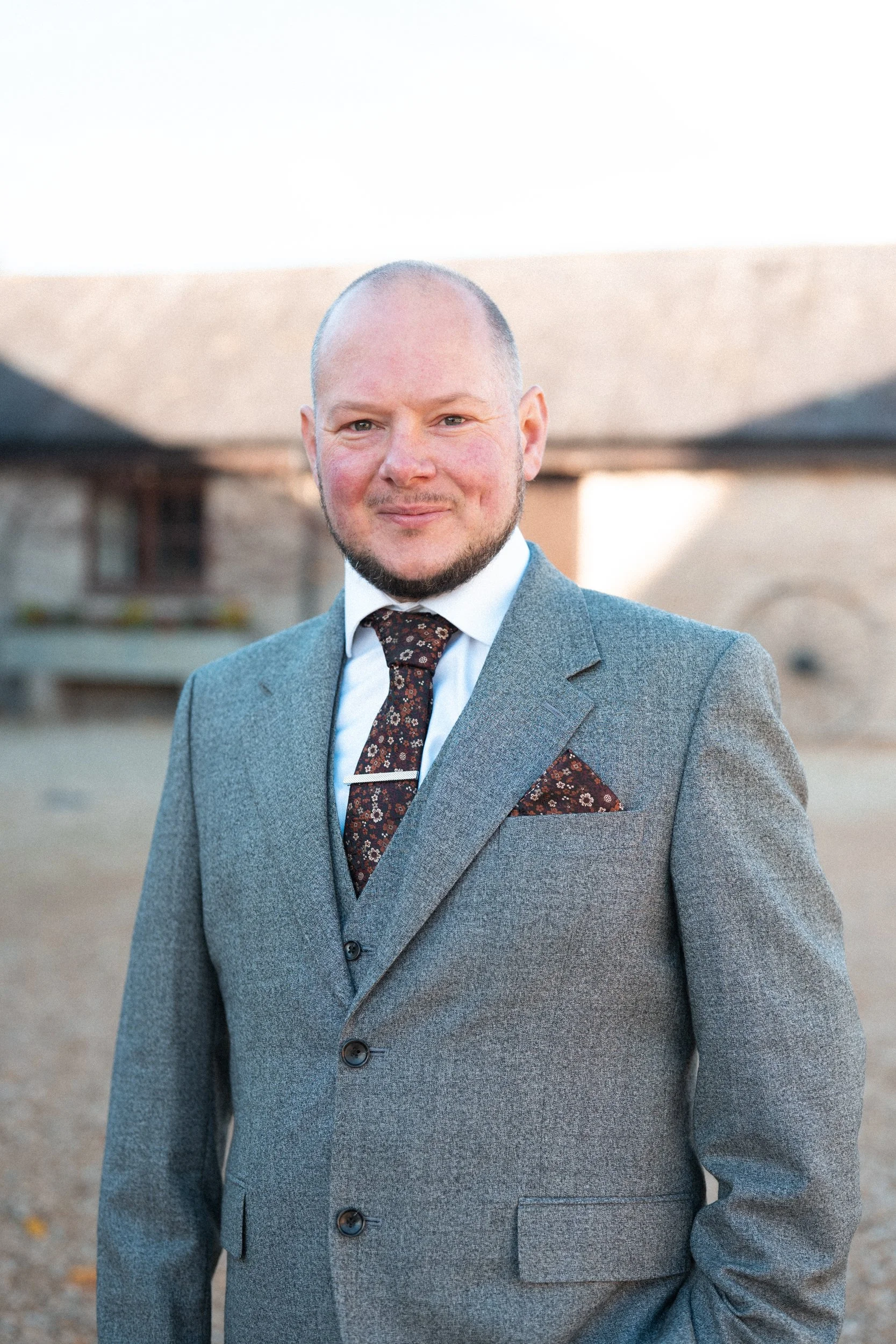 A smiling man in a gray suit with a floral tie and pocket square, standing outdoors with a blurred house in the background.