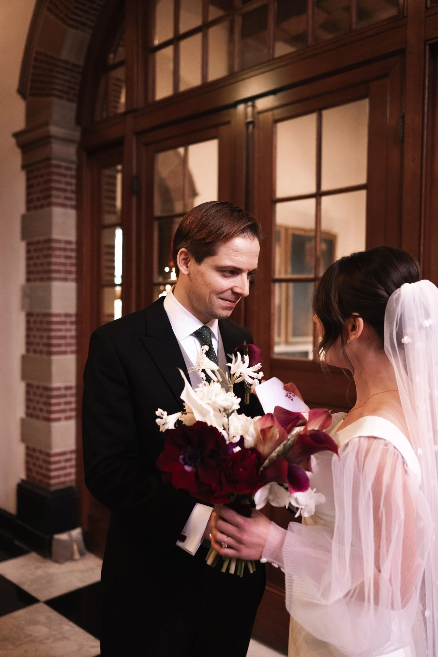 A groom in a black suit and a bride in a white wedding dress share a moment indoors with wooden framed glass doors and brick wall background, as he hands her a bouquet of red, white, and purple flowers.