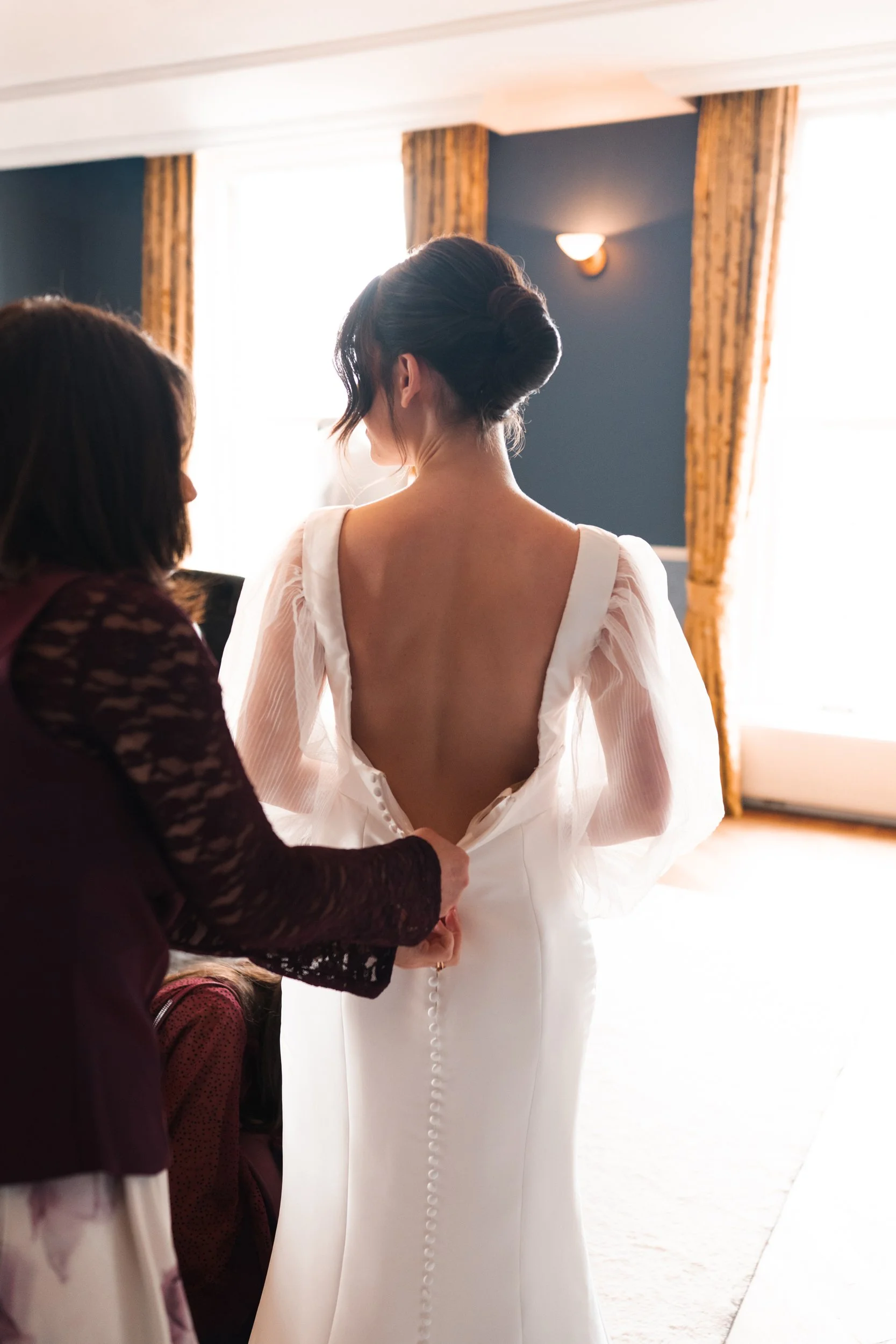A bride being assisted with her wedding dress by a woman, with sunlight coming through large windows and yellow curtains in the background.