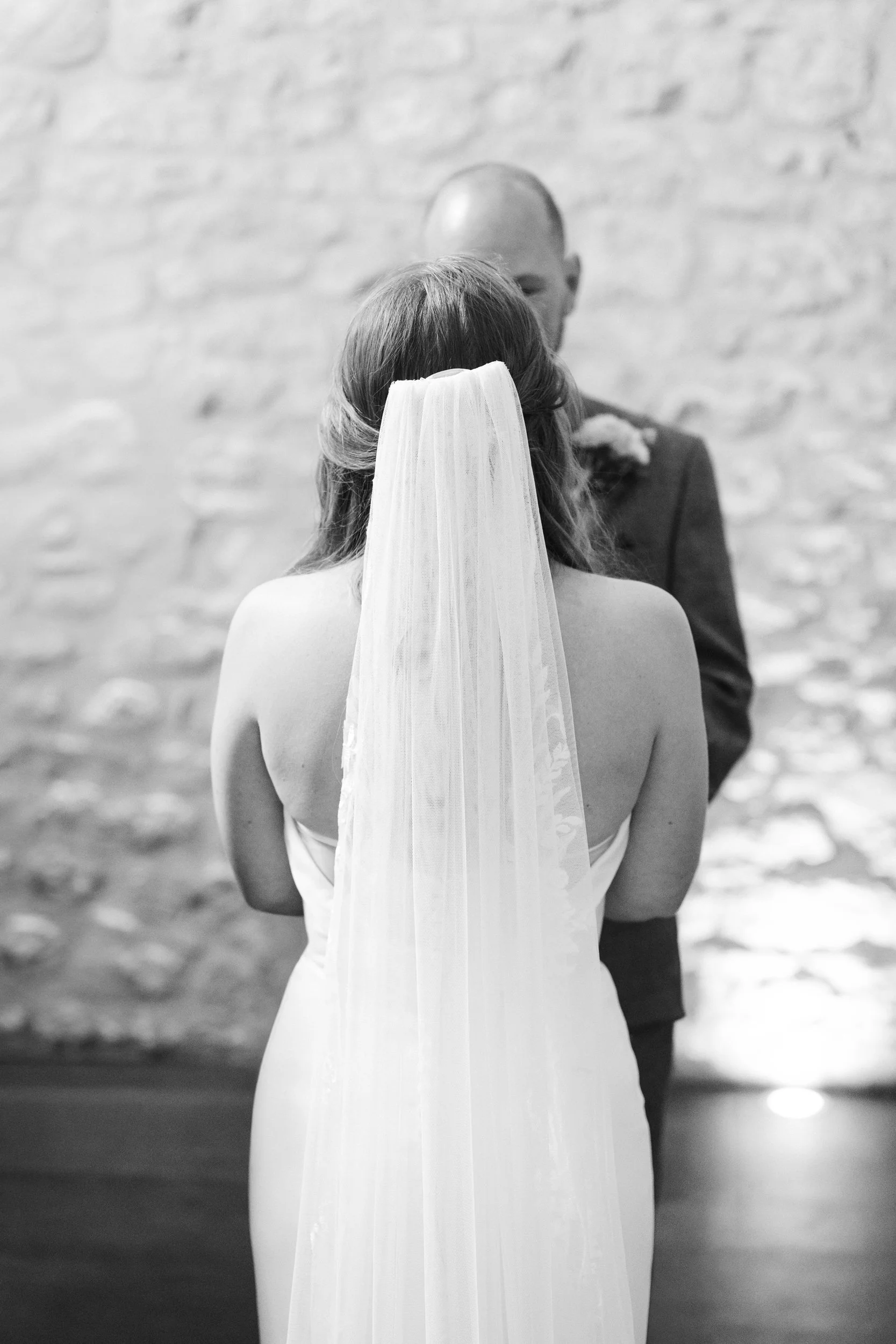 A bride with long hair wearing a veil and strapless gown faces a groom, who is bald and dressed in a suit, during a wedding ceremony against a brick wall background.