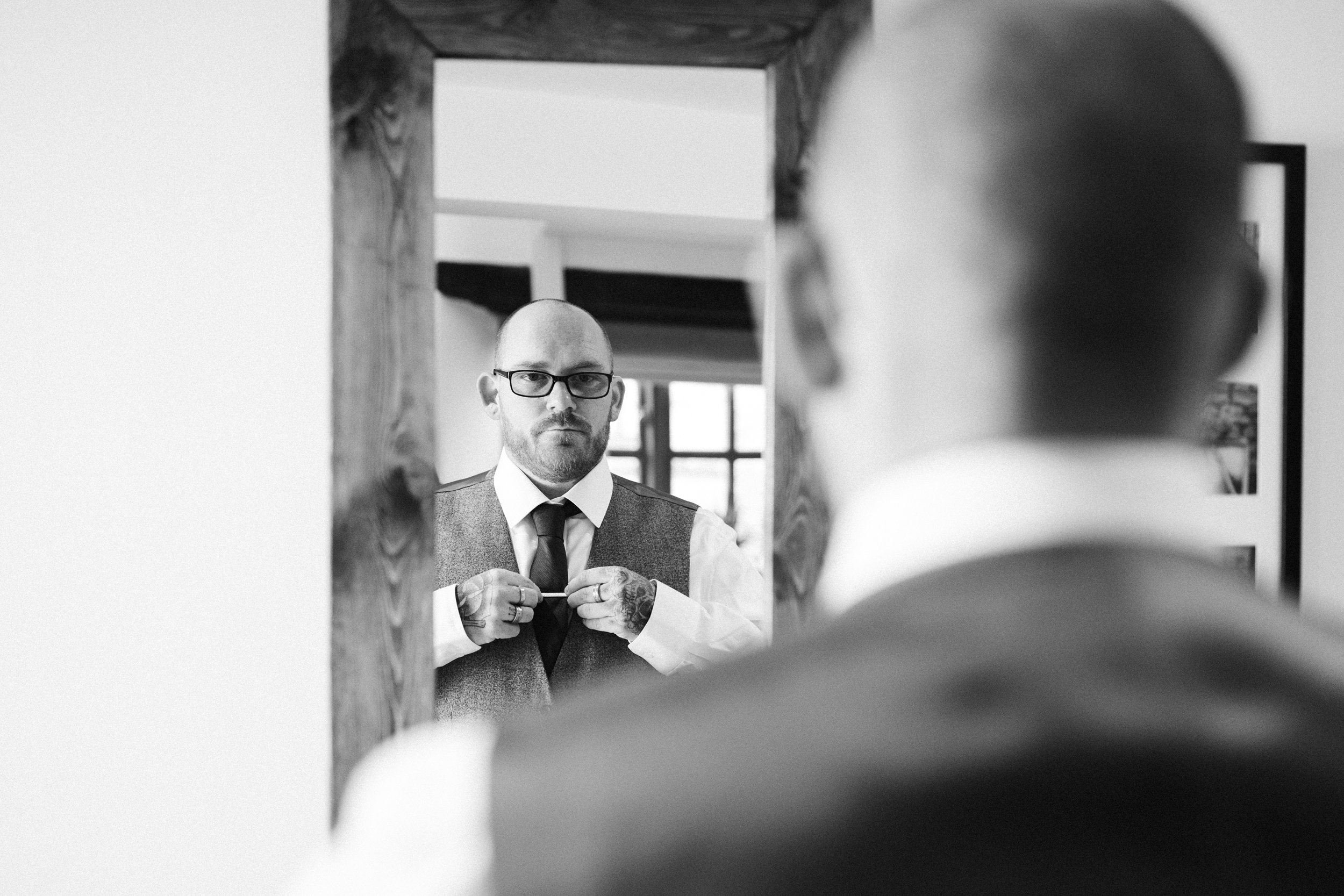 A man with glasses, a beard, and tattoos adjusts his tie while looking into a mirror. The reflection shows him wearing a formal vest and shirt, with a window and framed pictures on the wall behind him.