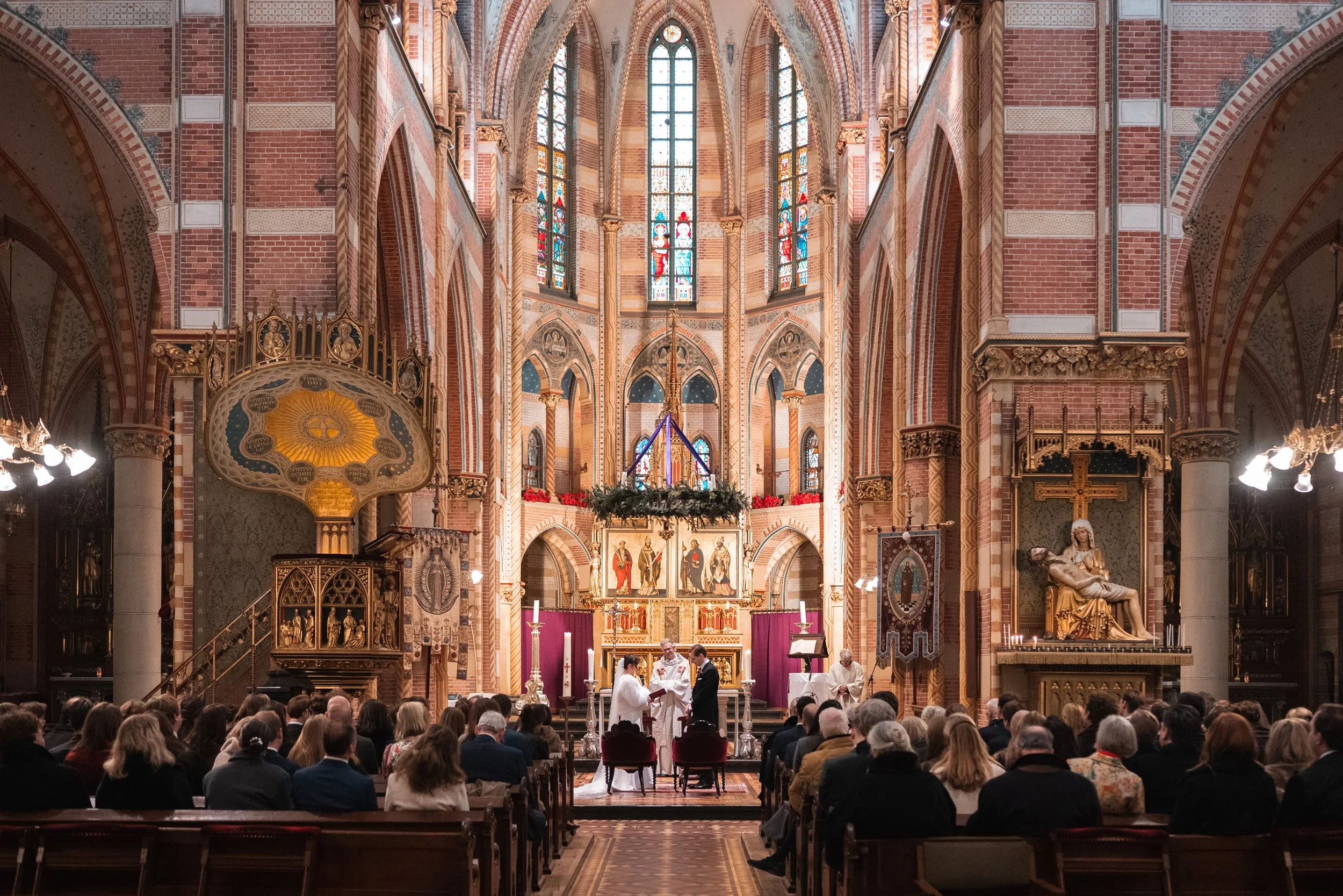A wedding ceremony taking place inside a large, ornate church with stained glass windows, as a priest officiates at the altar, and the bride and groom stand facing each other.