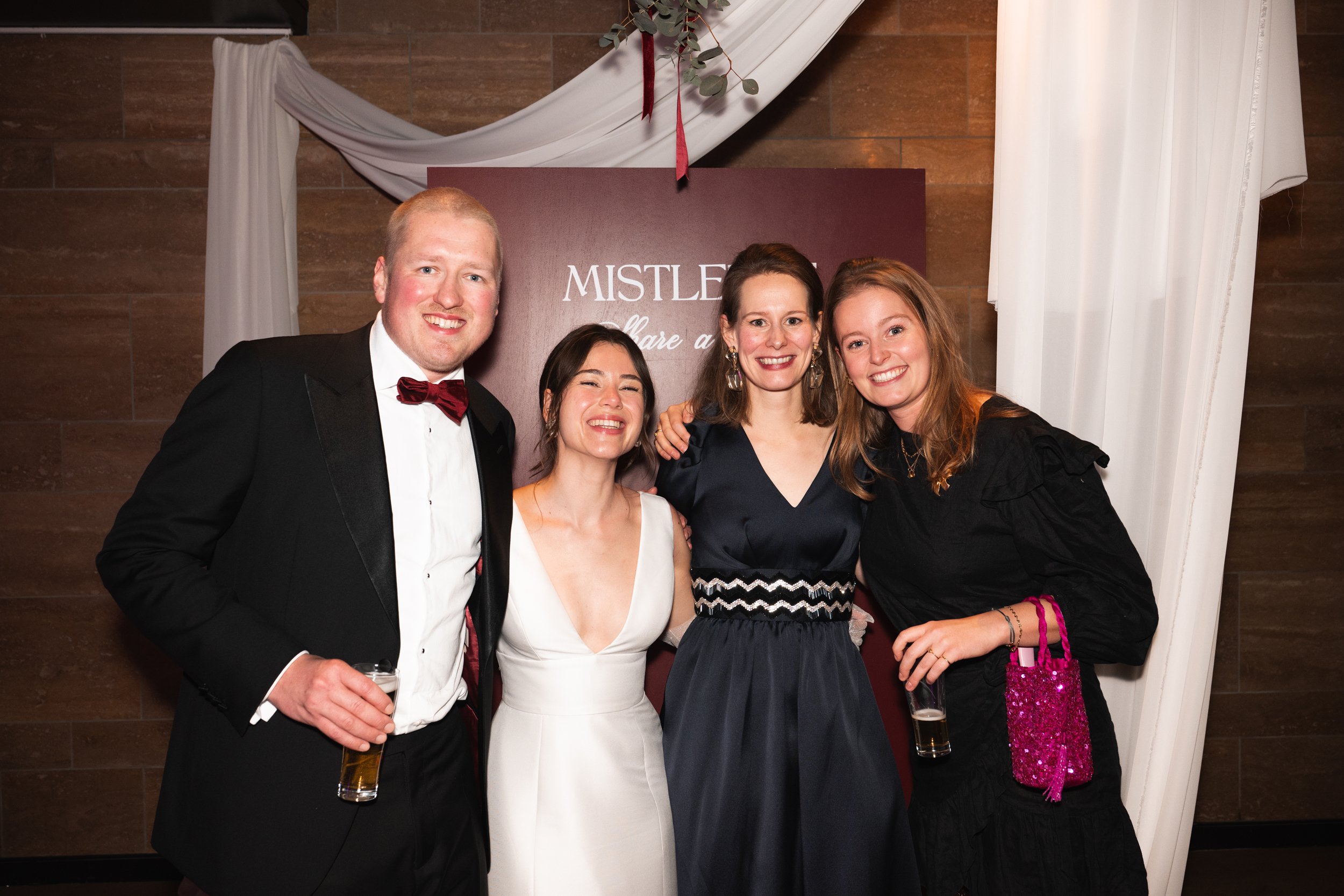 Four people dressed in formal attire, posing together and smiling at a celebration or event, with a sign reading 'MISTLE' in the background.