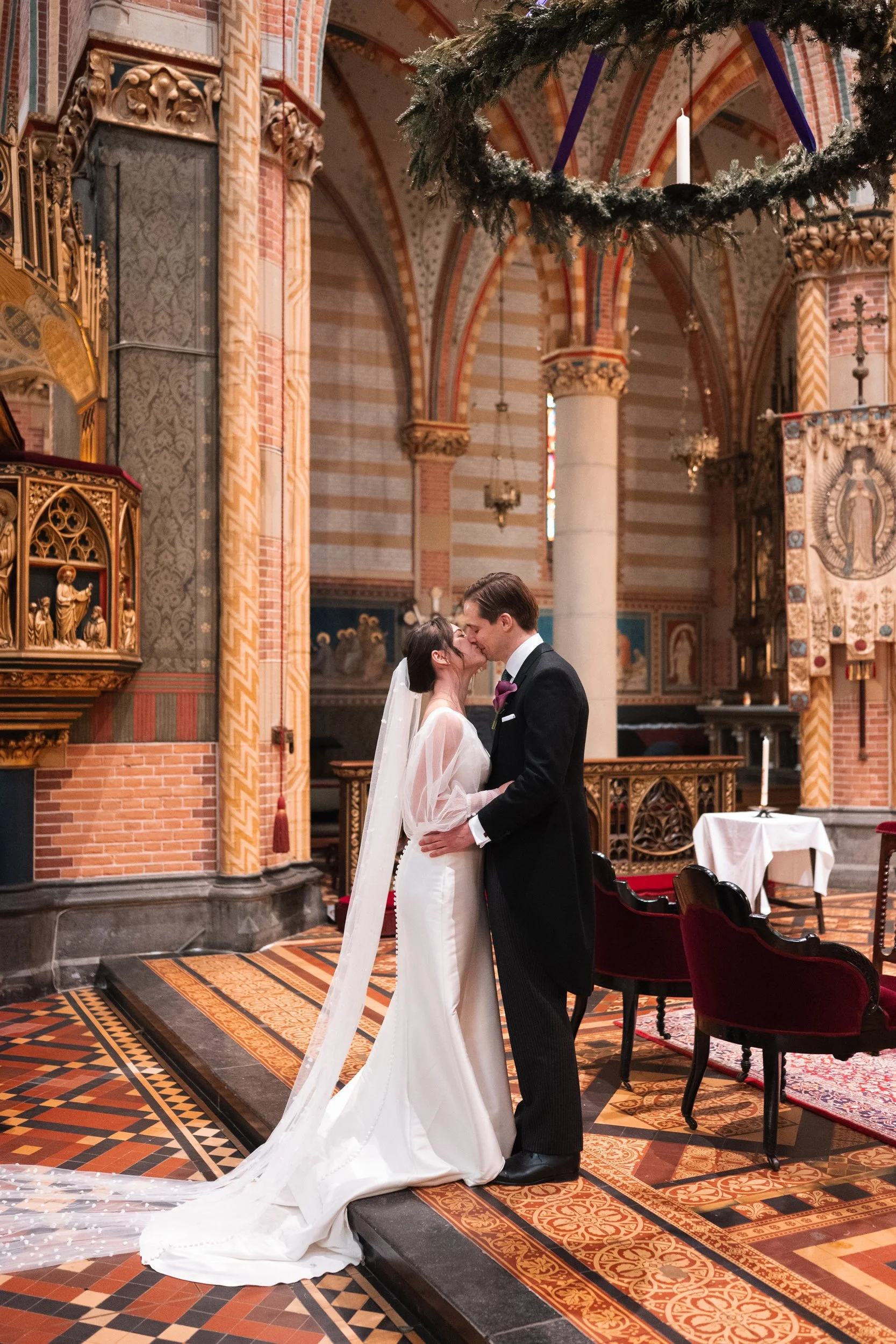 A bride and groom sharing a kiss inside a church, dressed in wedding attire with ornate decorations and stained glass windows in the background.