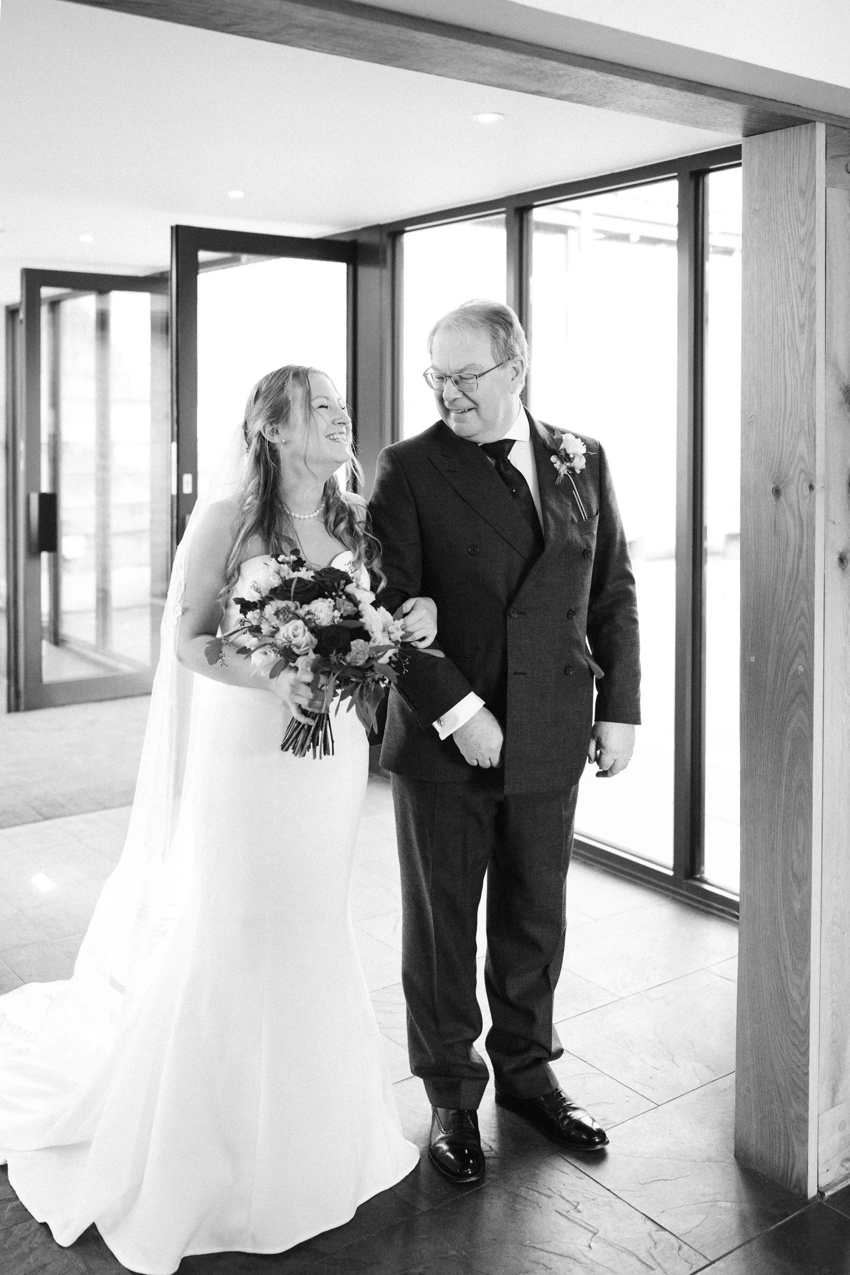 A bride smiling as she walks down the aisle with her father, holding a bouquet of flowers, in a wedding venue with large windows.