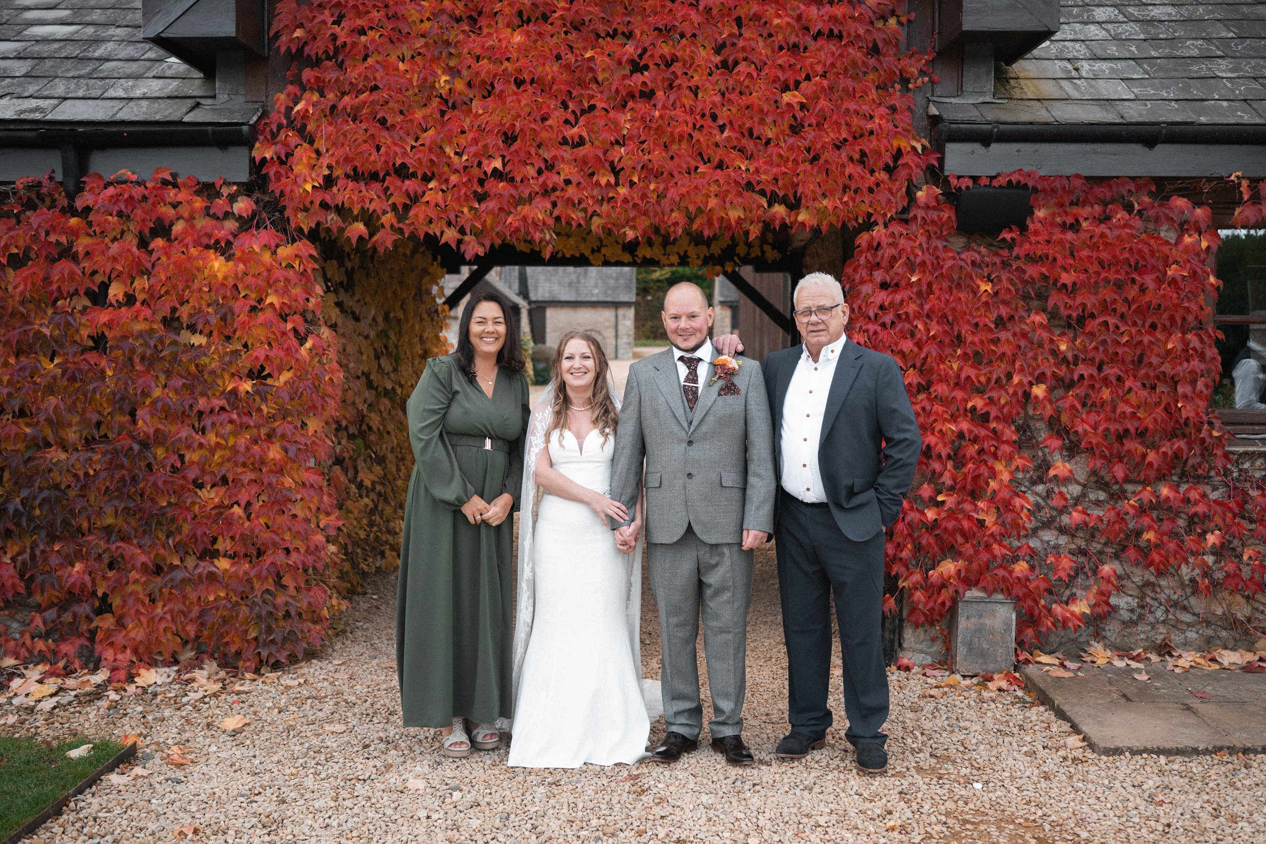 Group of five people at a wedding, standing under a wooden archway covered in red and orange autumn leaves. Two women and three men, one of whom is a bride in a white dress, and the others in formal attire, smiling for the camera.