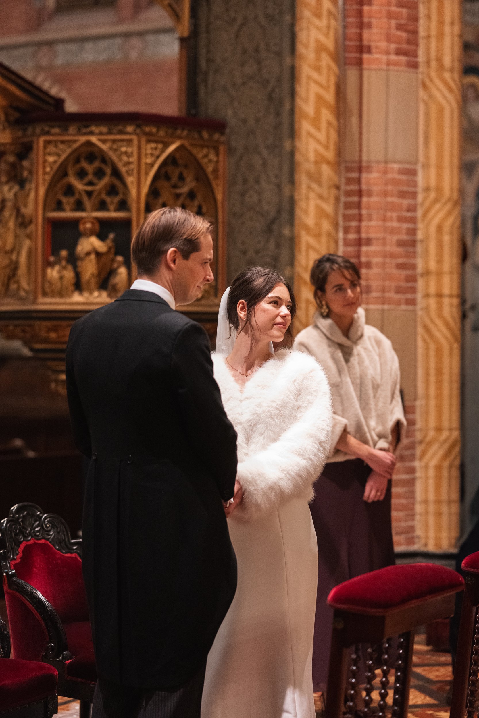 A bride and groom standing inside a church during their wedding ceremony. The bride wears a white gown and a furry white shawl, and the groom is dressed in a black tuxedo. A woman stands behind them, observing.