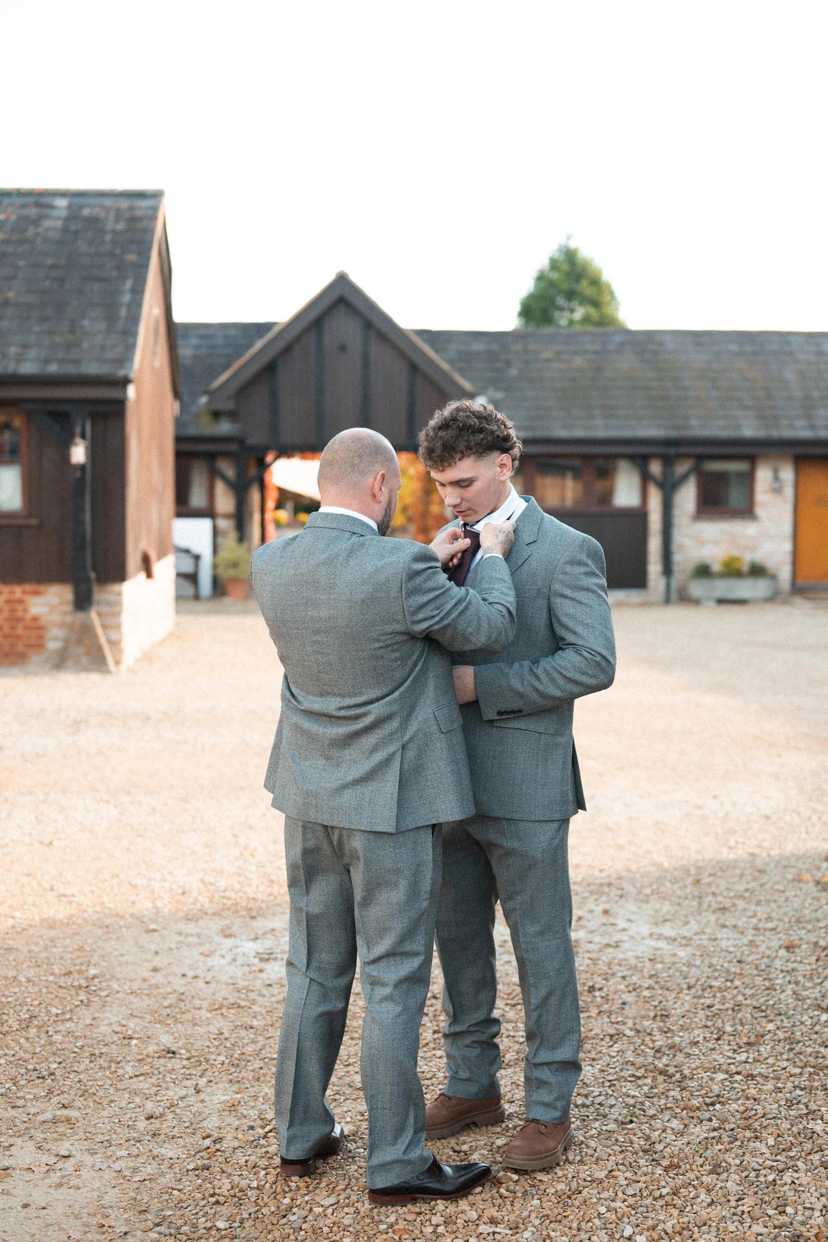 Two men in gray suits standing on a gravel surface outside a rustic building, one helping the other with his tie.