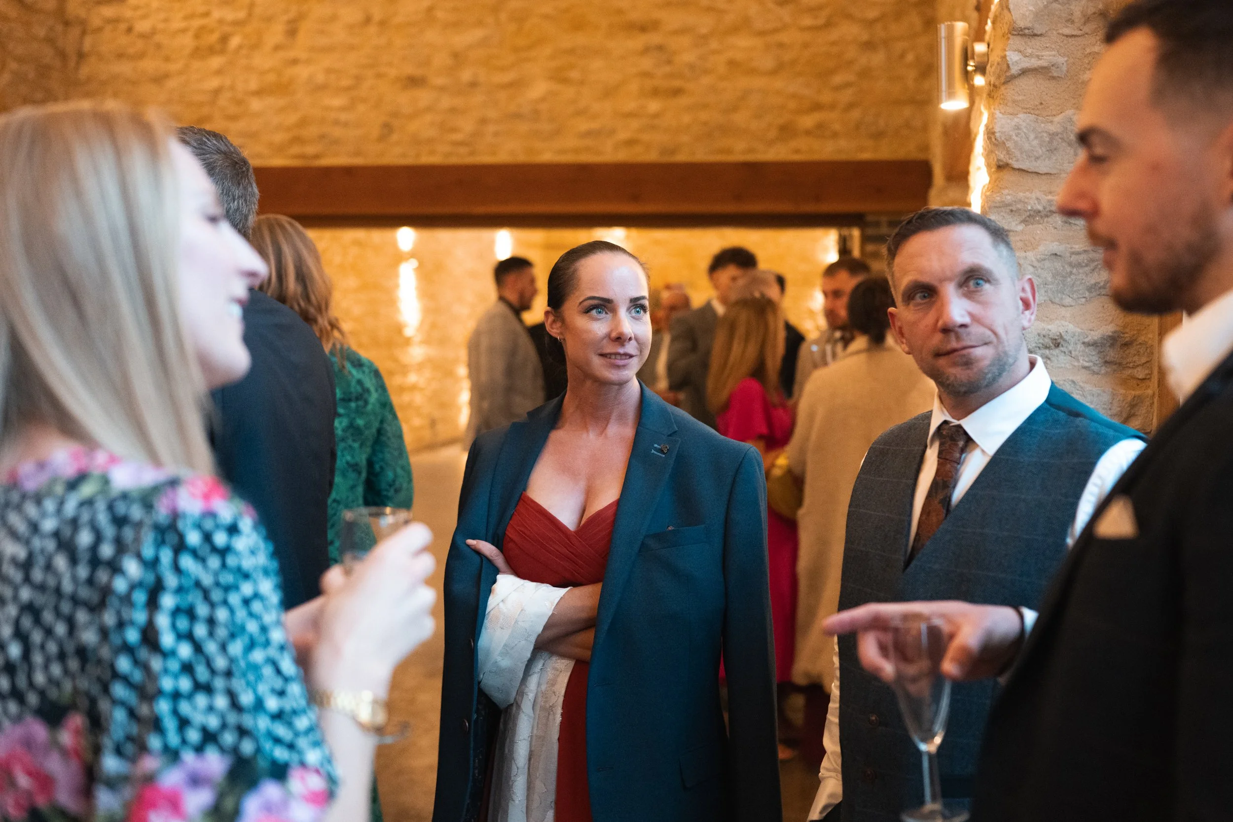 Group of people socializing at a party in a warm, rustic setting with stone walls, some holding drinks, engaging in conversation.
