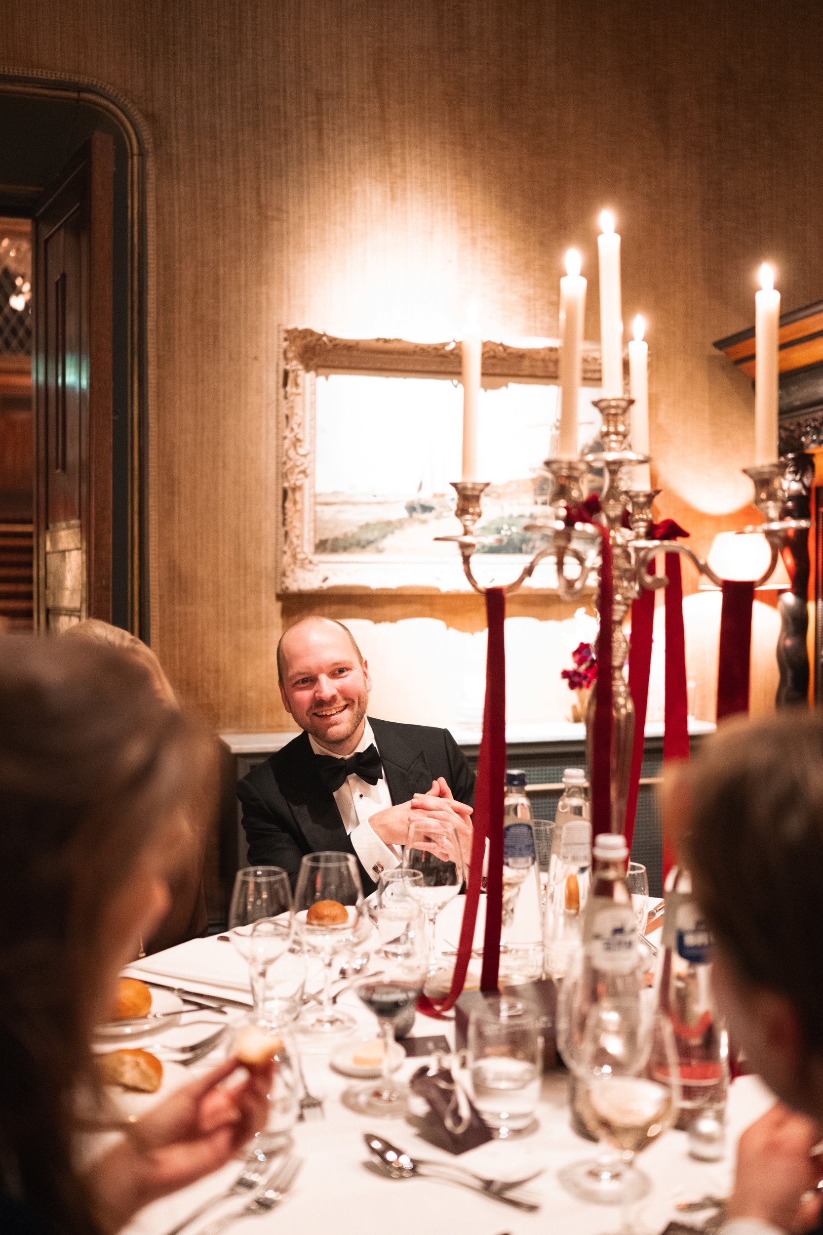 A man in a tuxedo smiling at a formal dinner event with multiple guests, set with wine glasses, plates, and bread rolls, and decorated with a silver candelabrum with white candles and red ribbons.
