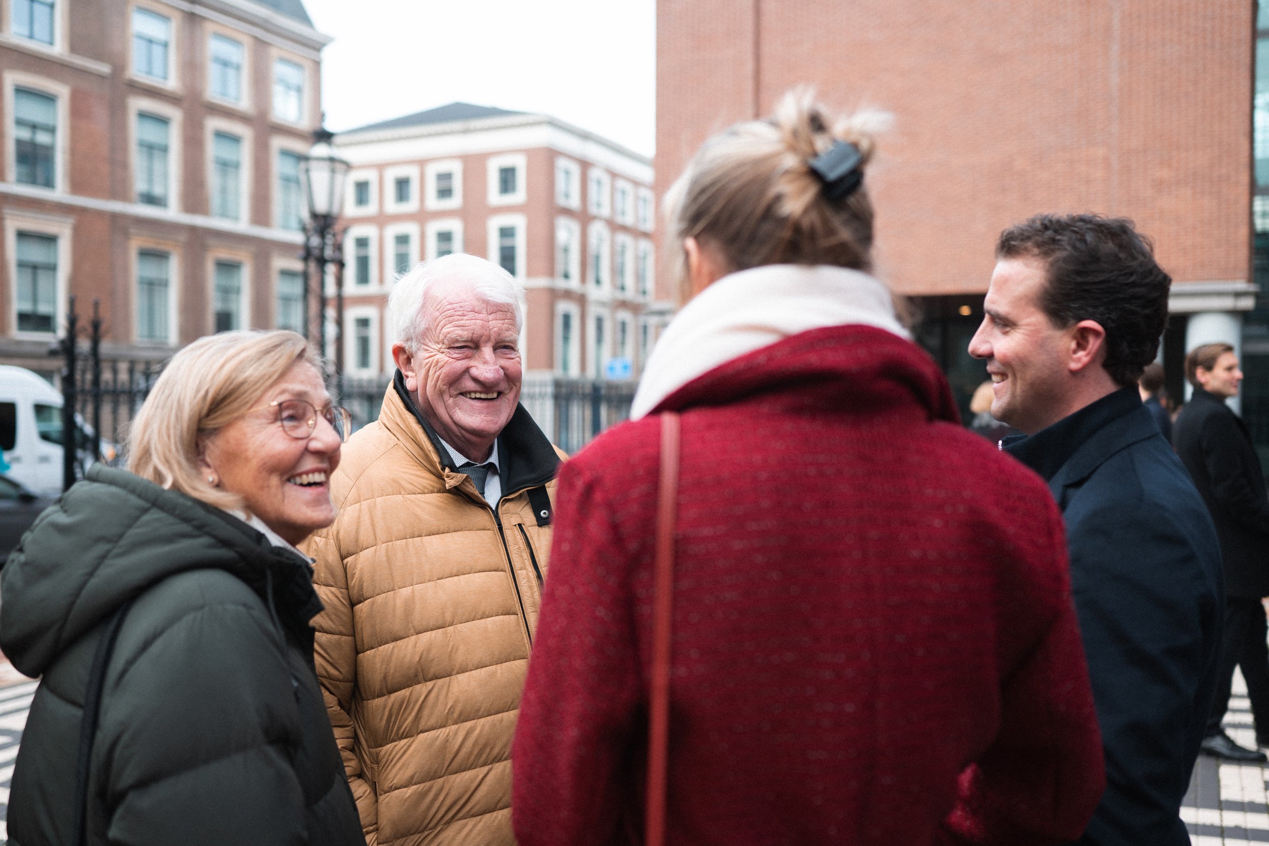 Four people smiling and talking outdoors in an urban area, with brick buildings and a street lamp in the background.