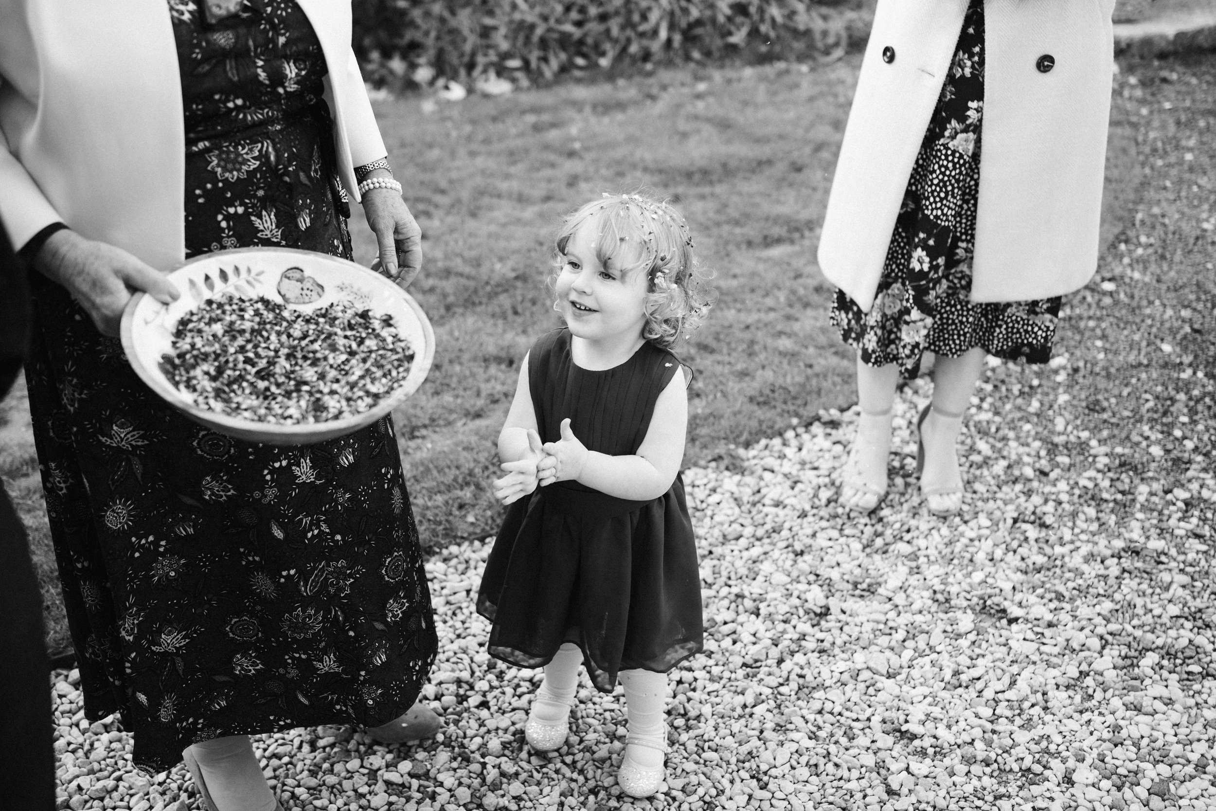 A young girl looking up and smiling at an outdoor gathering, surrounded by women dressed in floral and solid coats, with one woman holding a decorated tray. The scene is set on a gravel path with grass in the background.