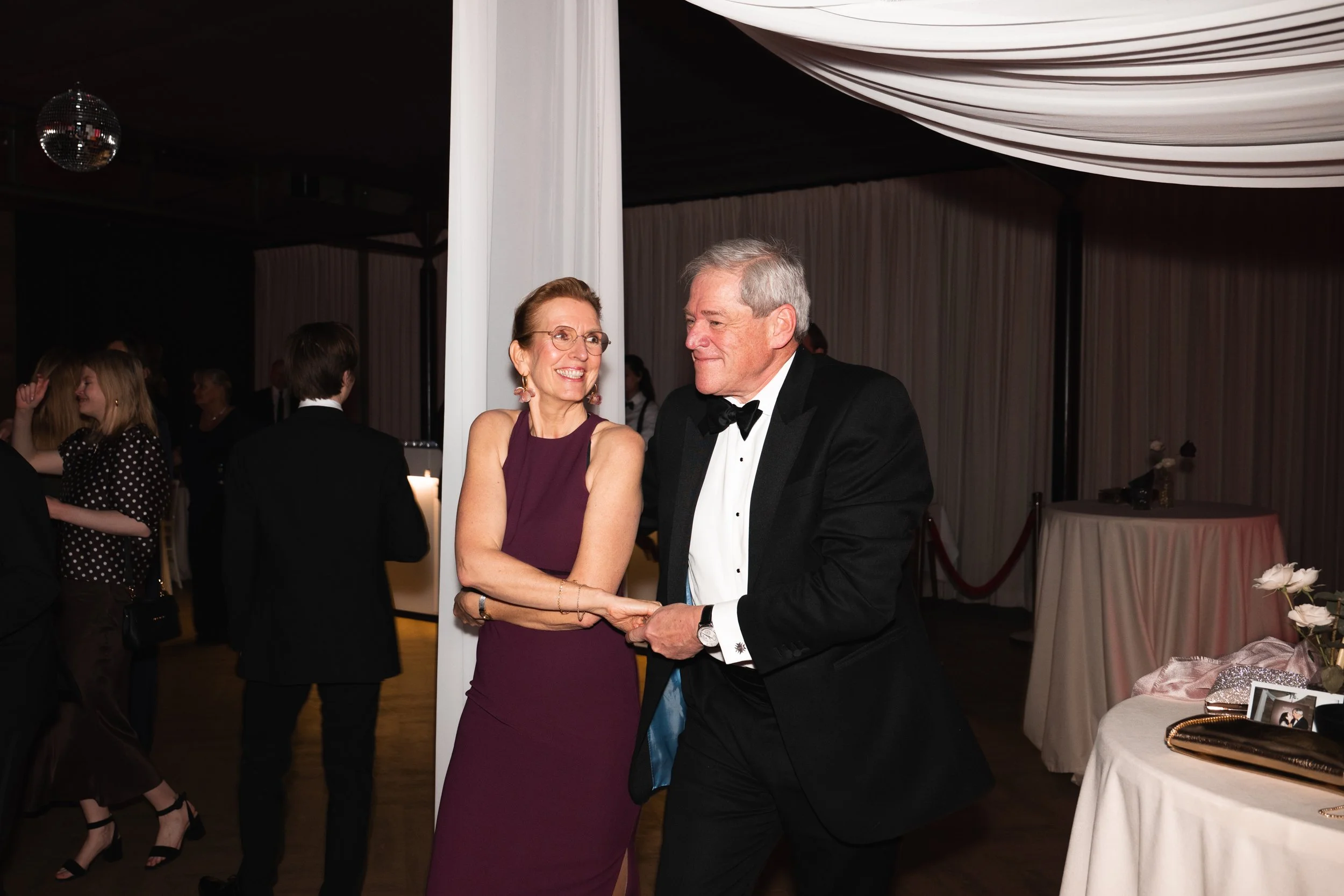 An elderly couple dancing at a formal event with other guests in the background.