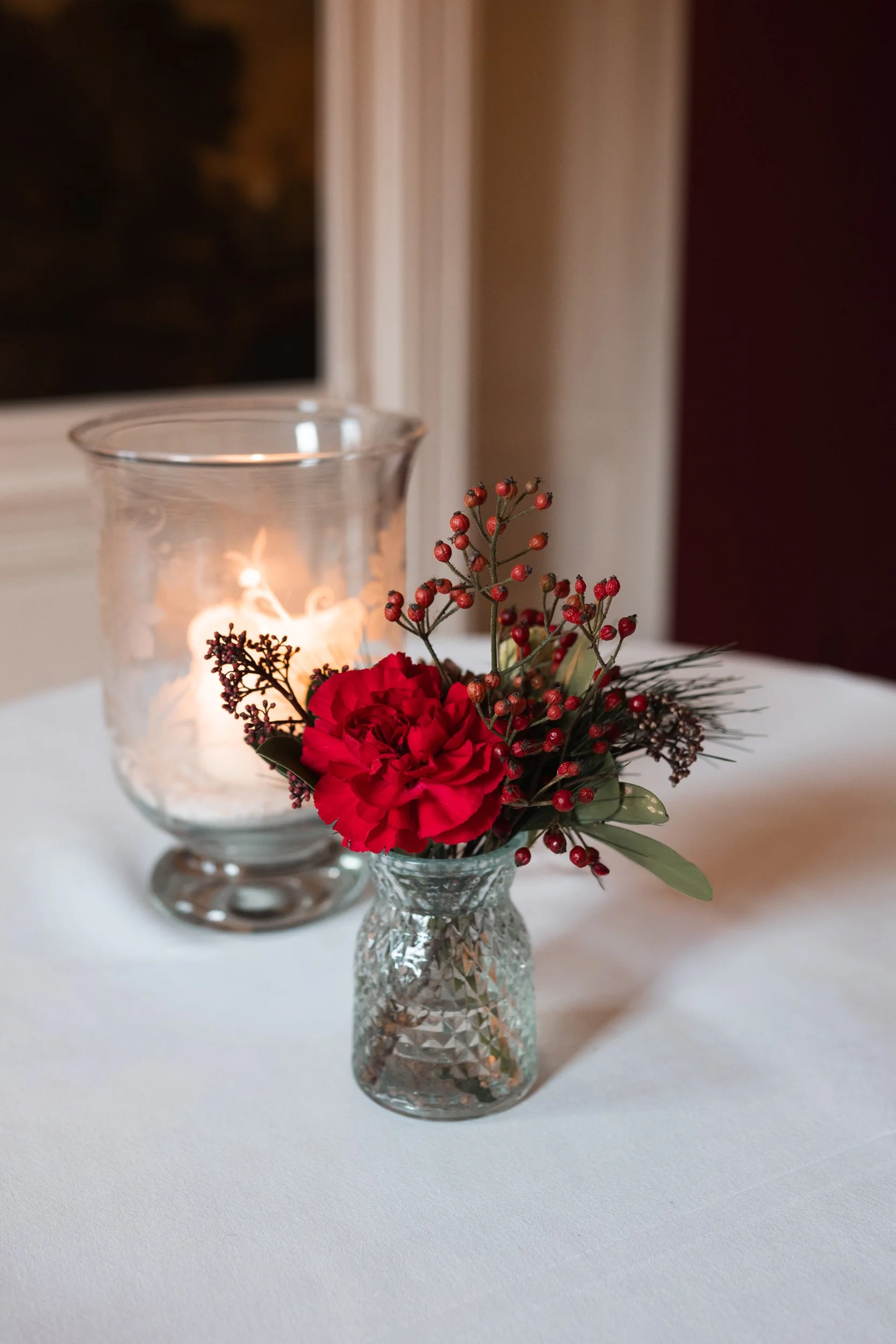 A small glass vase with red flowers and green leaves, placed on a white tablecloth, with a lit candle in a glass holder in the background.