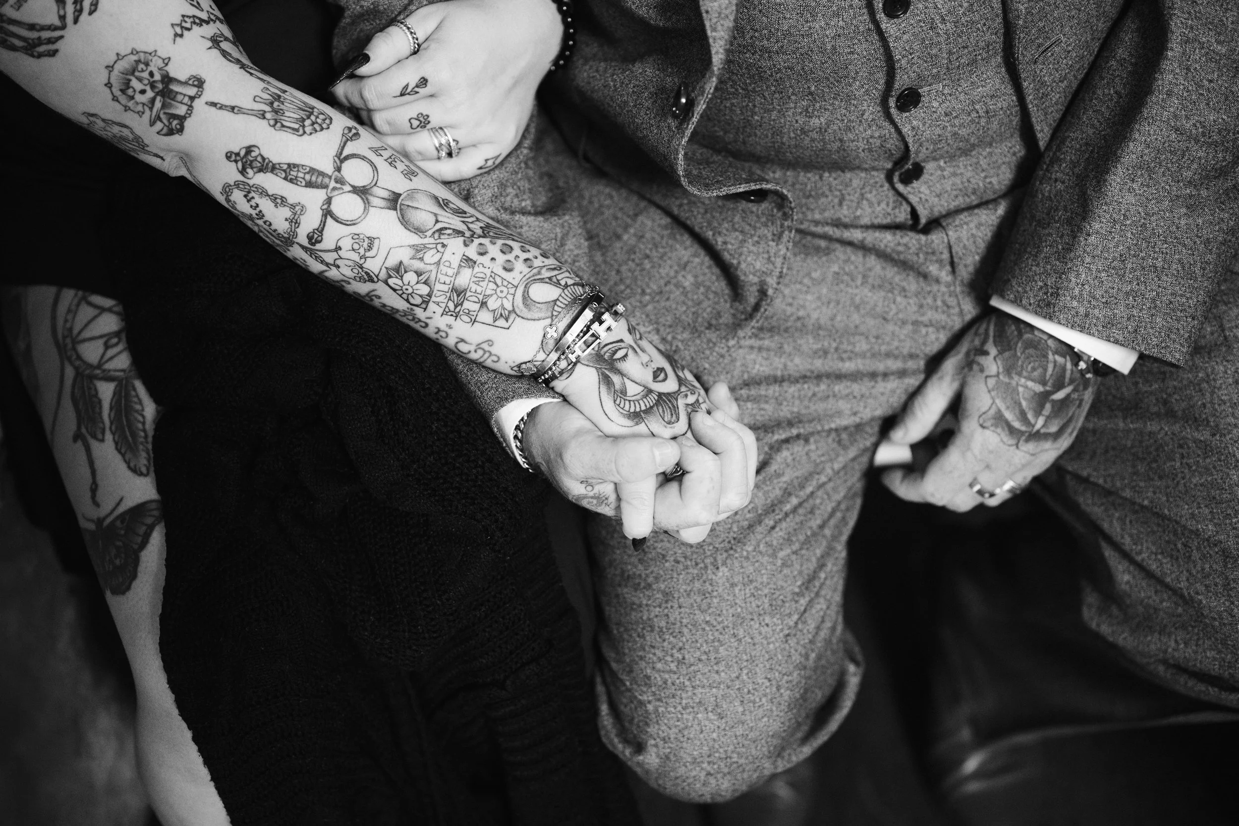 A close-up black and white photo of two people holding hands, showcasing tattoos on their arms and hands. One person is wearing a suit, and the other has colorful tattoos visible on their arm and hand.