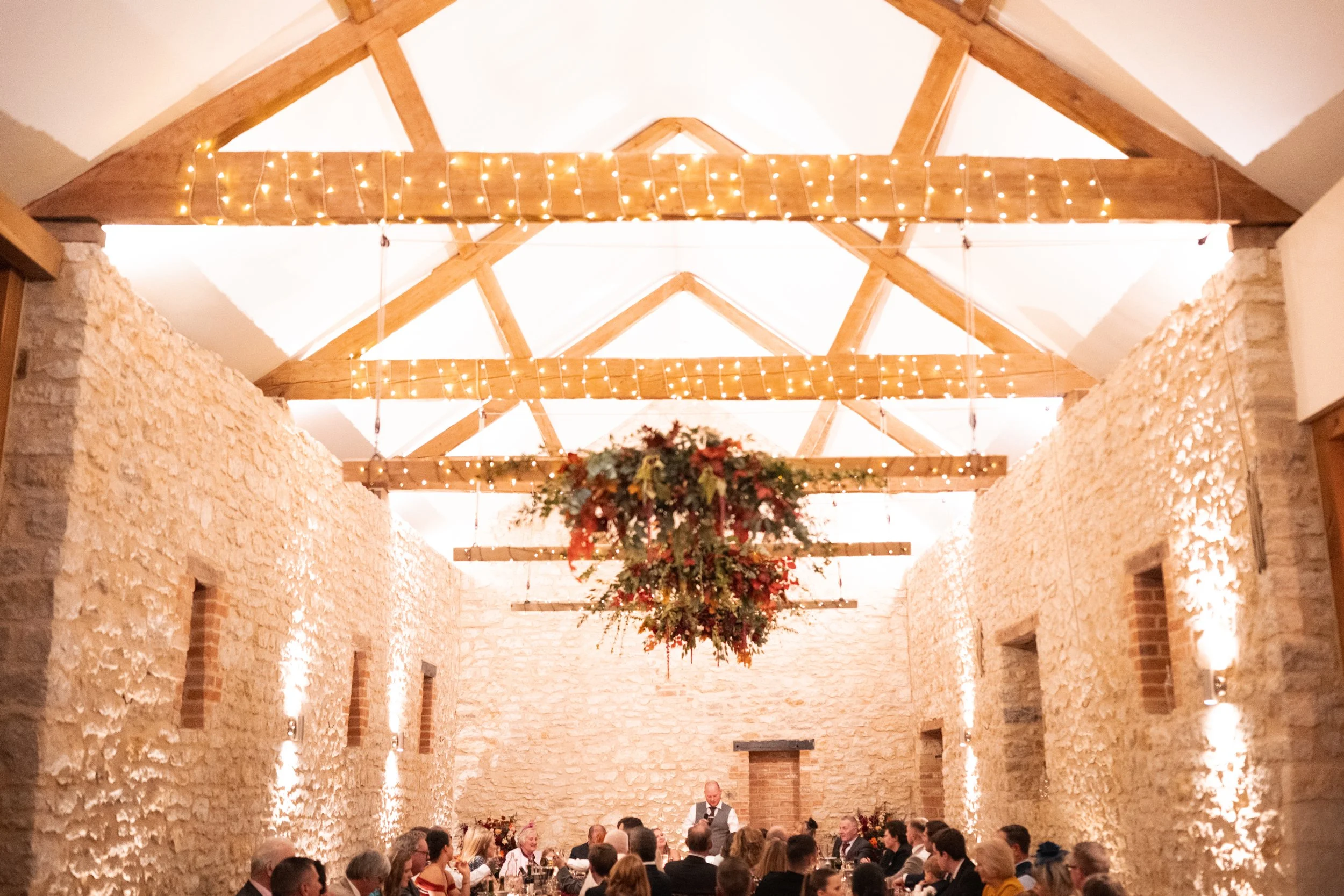 A large wedding or event reception hall with stone walls, wooden beams, and string lights on the ceiling. There's a floral chandelier hanging from the center of the ceiling. People are seated at long tables along the walls, and a person in a vest is 