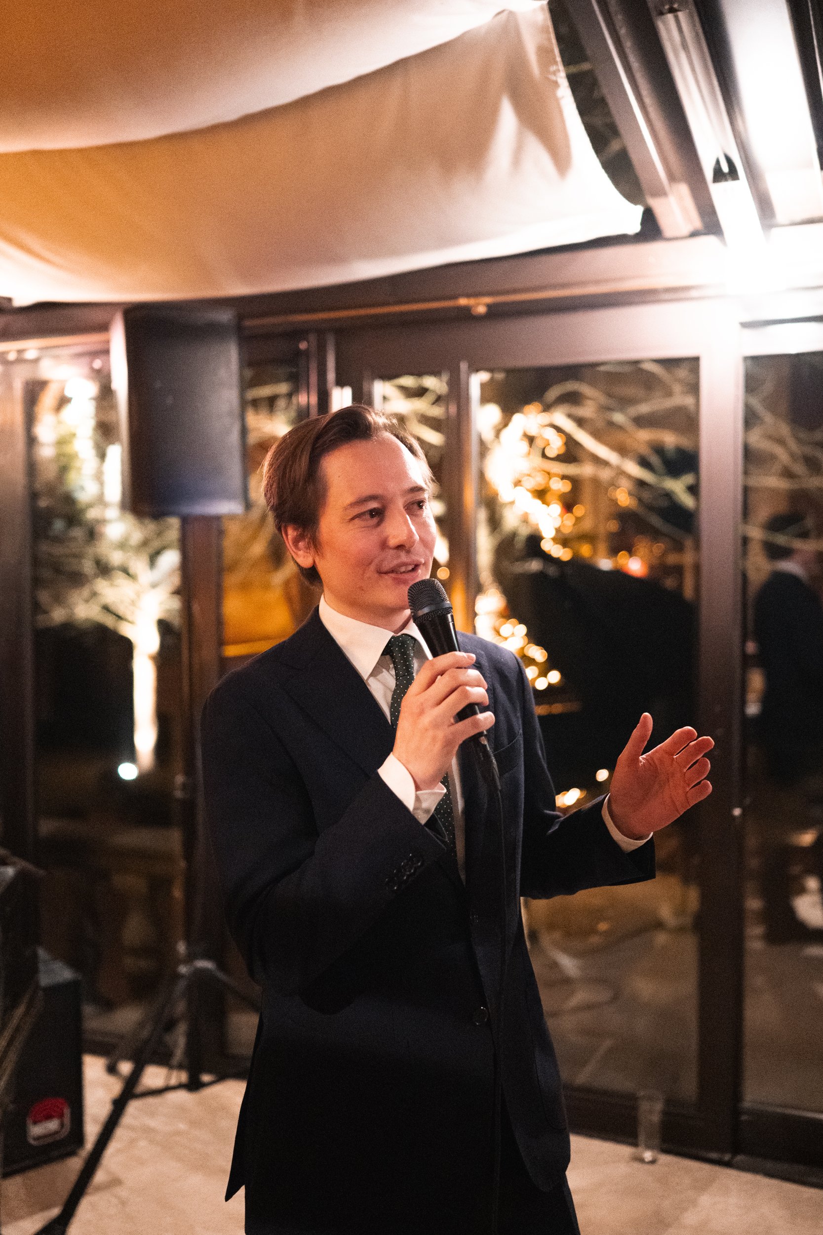A young man in a dark suit holding a microphone and speaking at an indoor event.