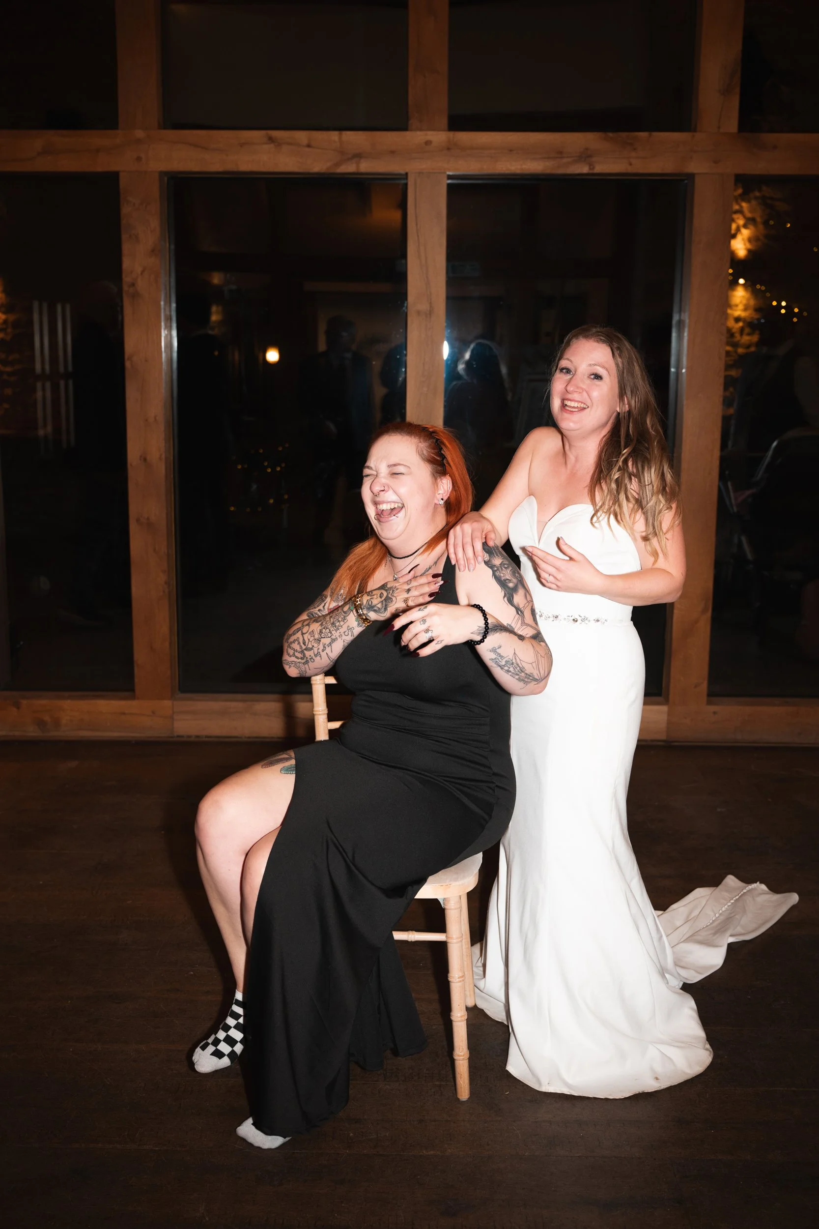 Two women at a wedding reception, one in a white wedding dress and the other in a black dress, laughing and enjoying themselves inside a venue with wooden walls and large windows at night.