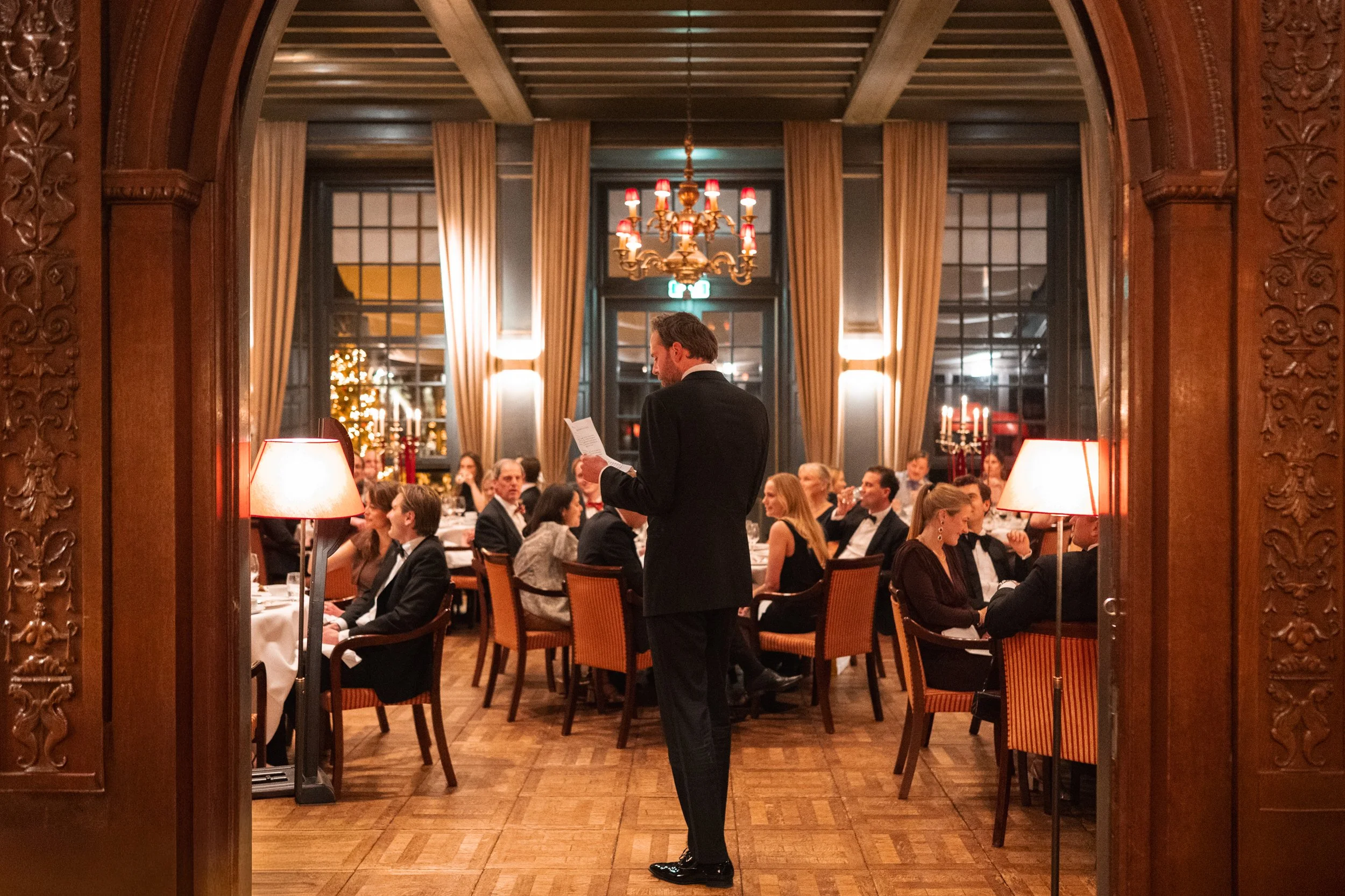 A waiter reads a menu to guests at a formal dinner in an elegant, warmly lit restaurant decorated with curtains, chandeliers, candelabras, and holiday decorations.