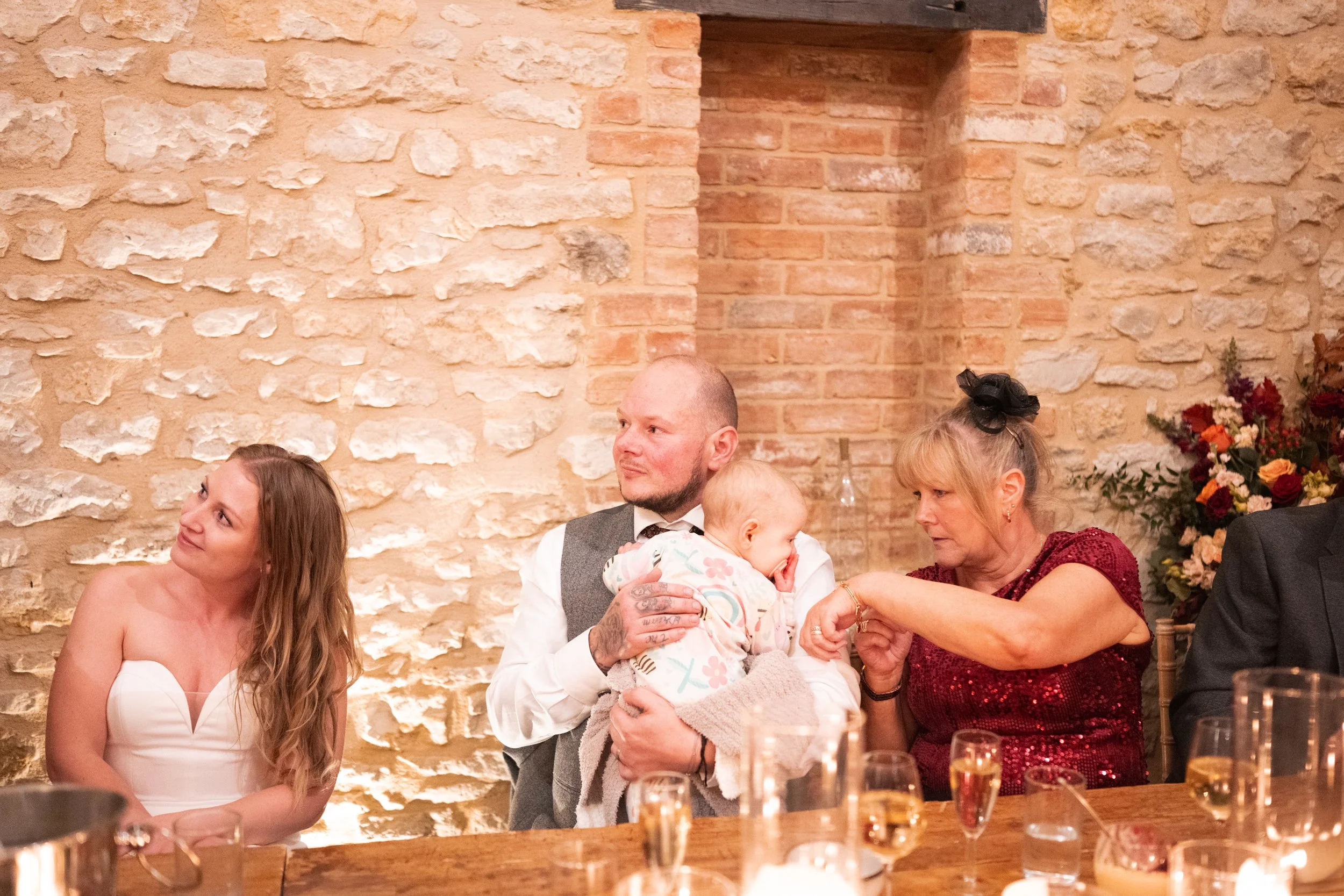 A group of people sitting at a table during a celebration, with a brick wall background, including a woman in a red dress and a man holding a child, and a young woman in a white dress on the left.