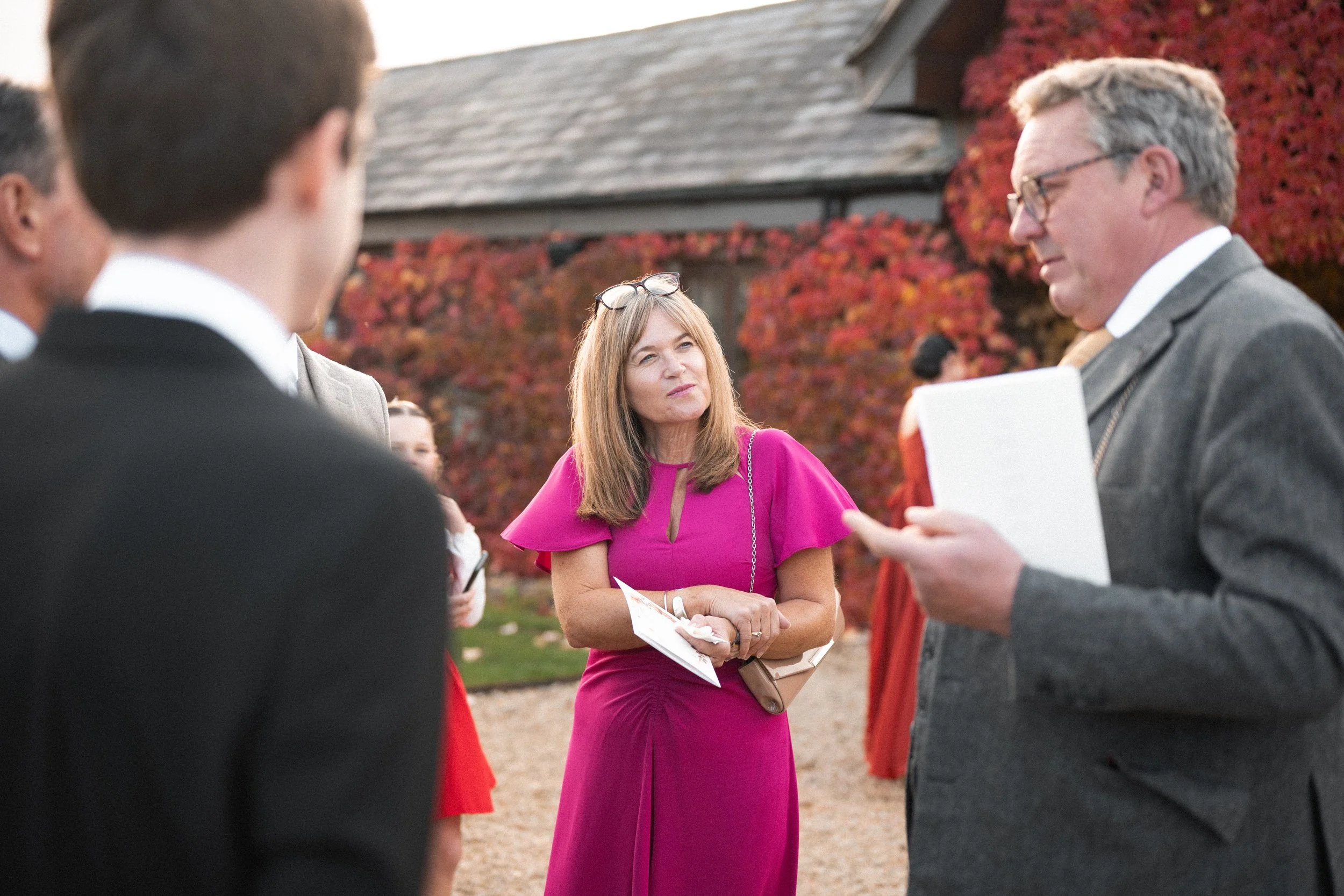 A woman in a bright pink dress is standing outdoors, listening to a man holding a document, while several other people are gathered around in conversation. There are autumn trees with red leaves in the background.