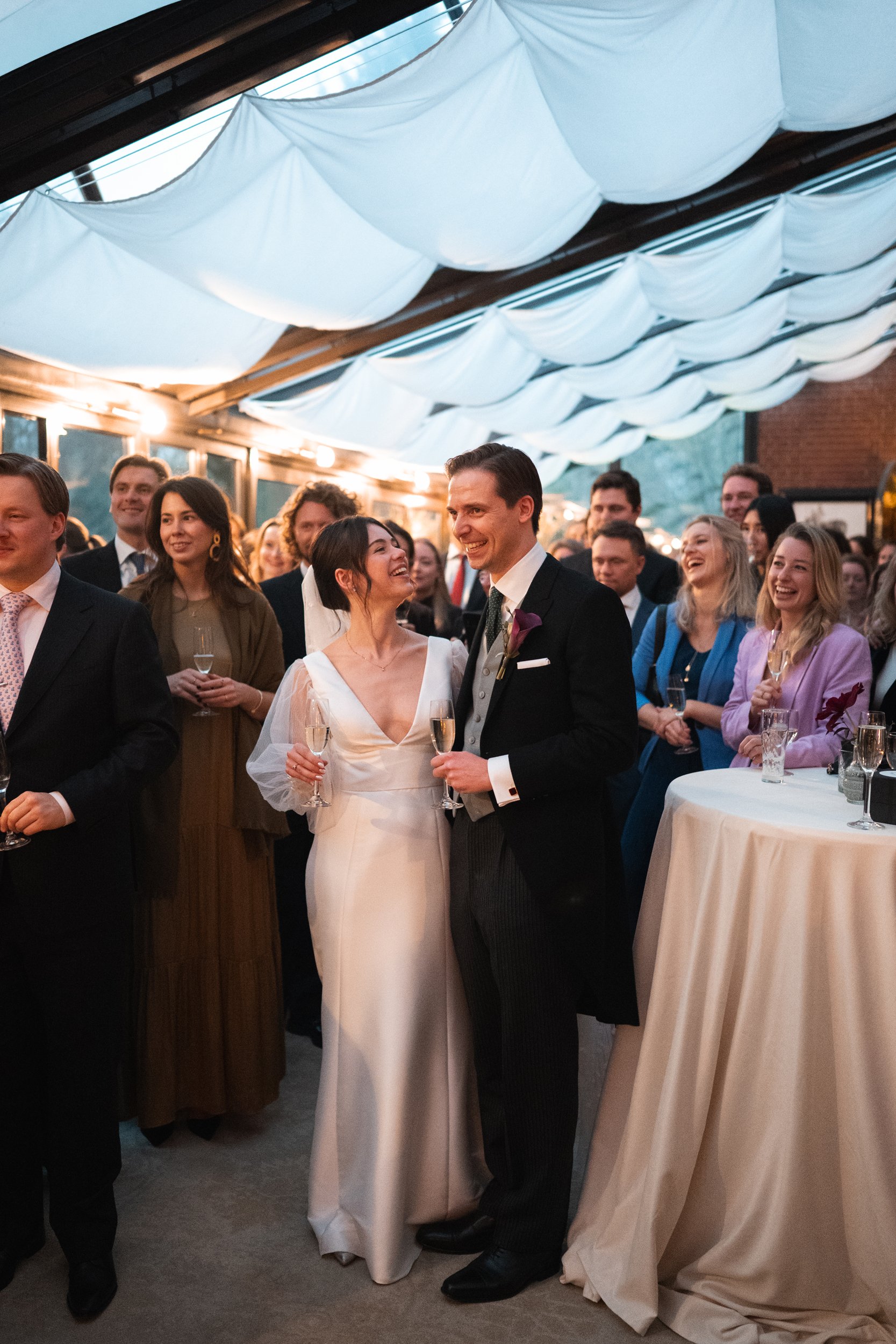 A bride and groom stand together at their wedding reception, smiling and holding glasses of champagne, surrounded by guests under a decorated ceiling with white fabric draping.