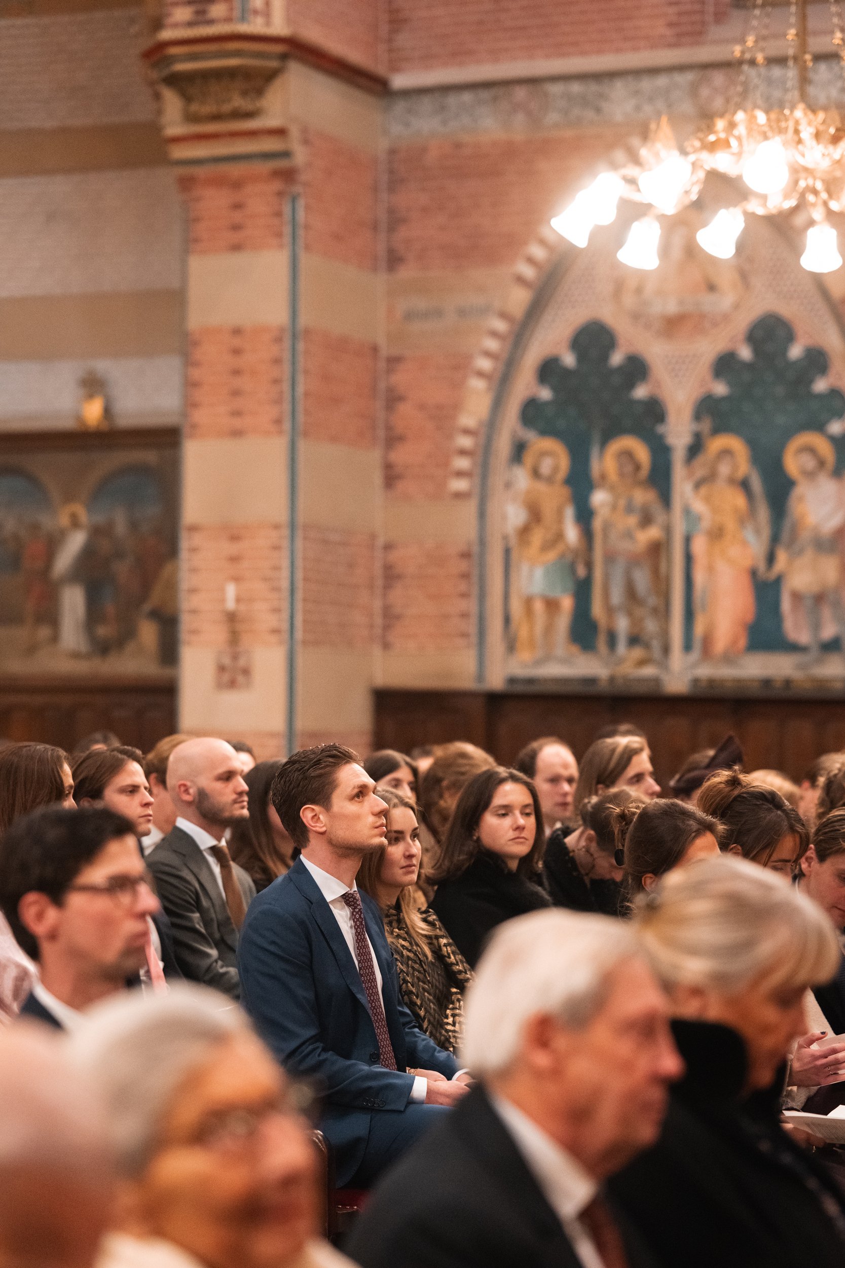 A group of people sitting attentively in a church or cathedral, with brick walls, religious artwork, and chandeliers in the background.
